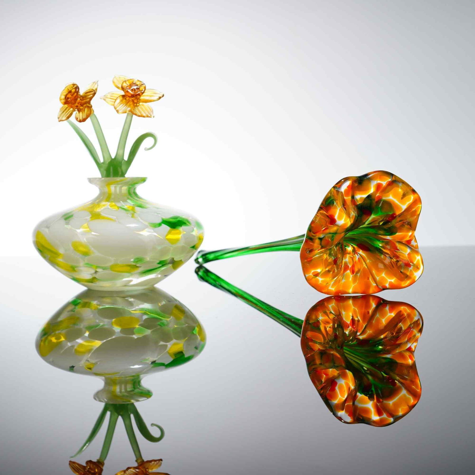 2pc Floral Motif Art Glass Bottle and Figure, Caesars World Daffodil Bottle and Orange Yellow Glass: This two-piece art glass grouping comprises a decorative figural bottle with stopper and a hand-blown floral sculpture. The bottle features a small, bulbous clear glass form with internal white, yello