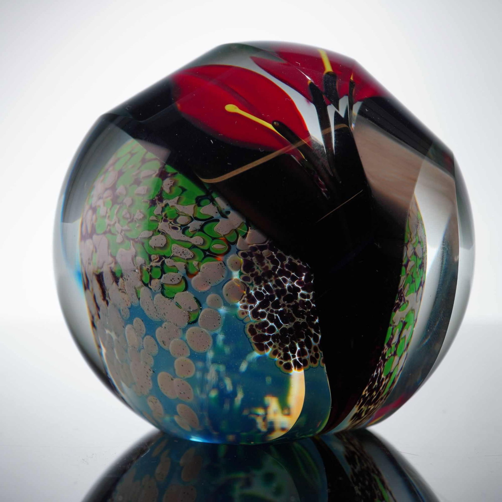Satara Faceted Art Glass Lily Paperweight: The interior features a three-dimensional lampworked red lily with yellow stamens and a dark stem. The floral motif is positioned over a mottled ground of green, blue, and brown frit inclusions, resem