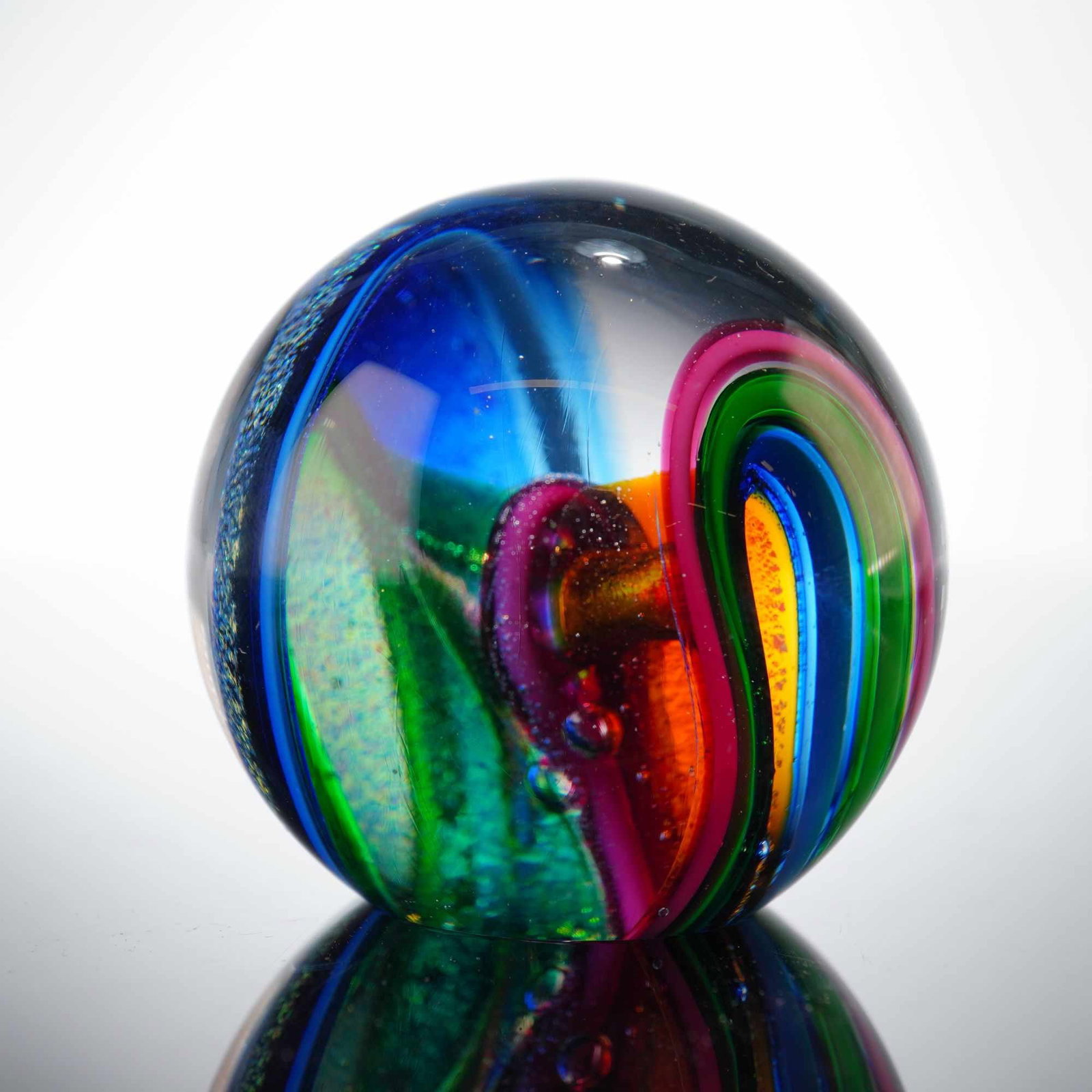 Glass Eye Studio Art Glass Paperweight: The spherical object is composed of a clear glass casing that encapsulates an internal abstract design featuring swirling ribbons of cobalt blue, emerald, green, magenta, and orange. The central core 
