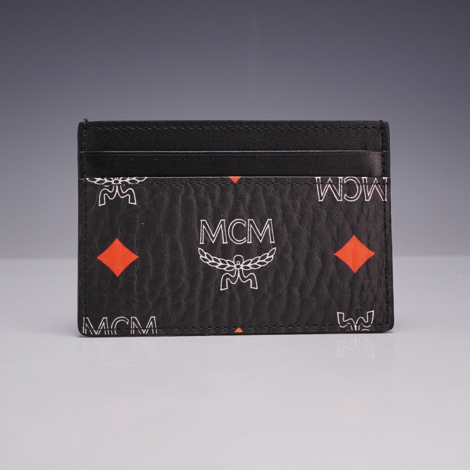 MCM Spectrum Diamond Visetos Black Leather Credit Card Holder with Original Box and Dust Bag: This MCM credit card holder is crafted from black pebbled leather featuring the Spectrum Diamond Visetos monogram print. The design consists of white outlined brand logos and laurel wreaths with solid