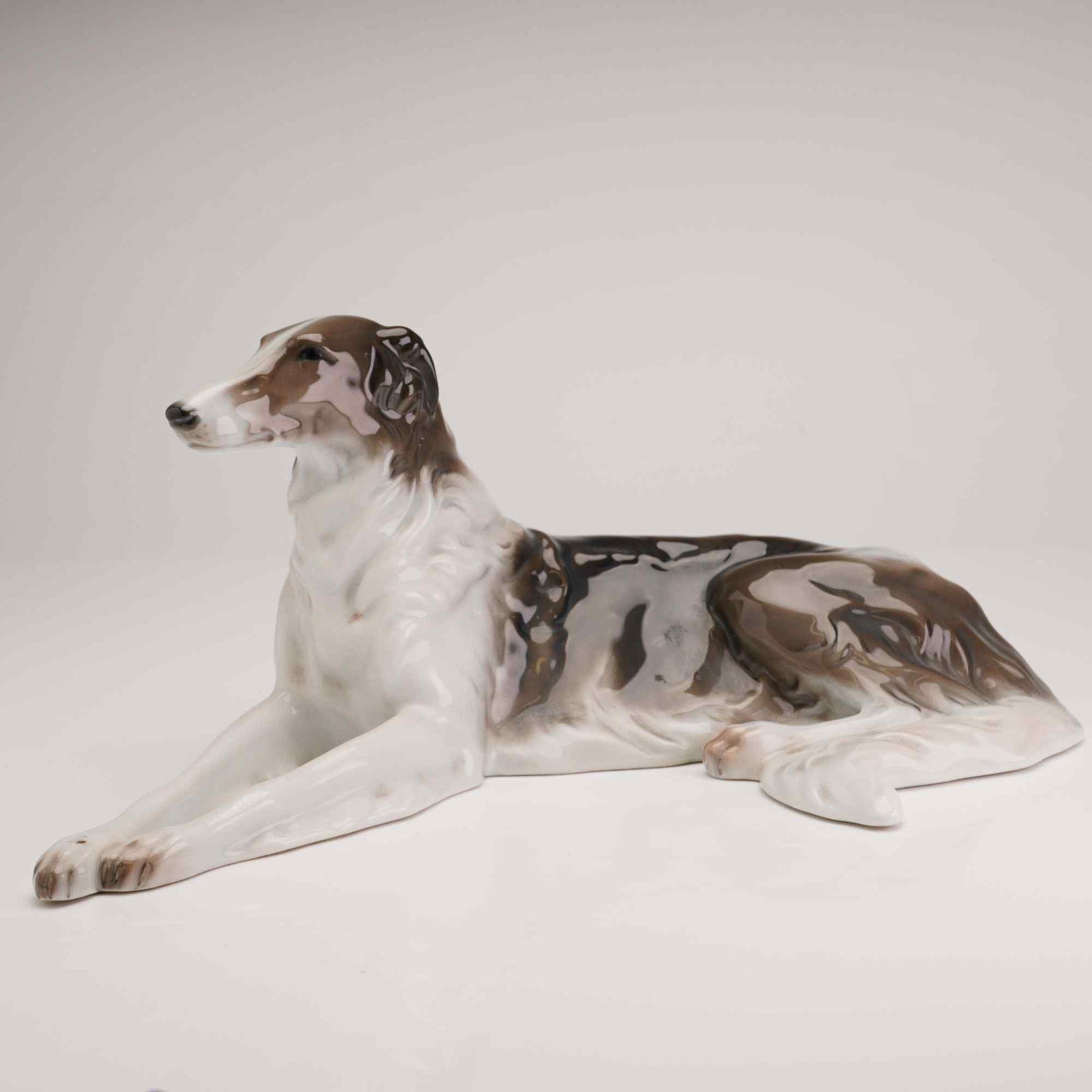 Rosenthal Porcelain Figurine of a Reclining Borzoi: A Rosenthal glazed porcelain figurine of a reclining Borzoi dog is modeled in a resting pose with extended front legs and the head turned slightly to the left. The piece features a high-gloss glaze wi