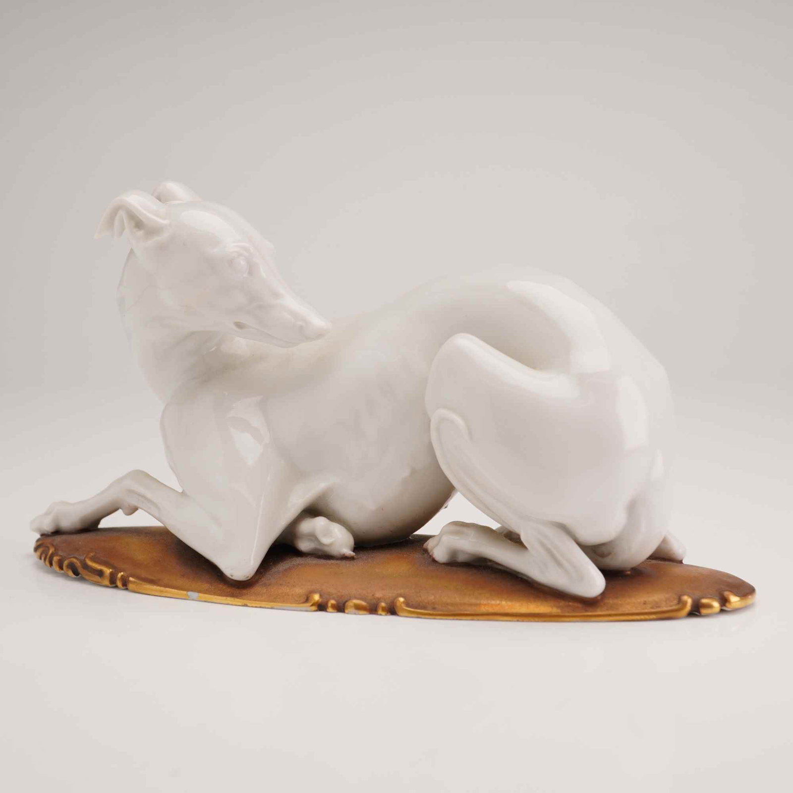 Rosenthal Porcelain Recumbent Greyhound Figurine: A glazed white porcelain figurine of a reclining greyhound or whippet, designed by artist Theodor Karner. The figure is modeled in a recumbent posture with the head turned toward the flank, resting on