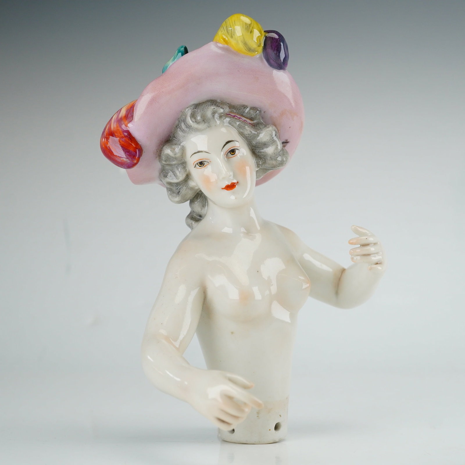 Porcelain Half Doll with Four-Plumed Bonnet: This hand-painted glazed porcelain half doll depicts a woman from the waist up, modeled with gray-powdered hair in curls. She wears a large, wide-brimmed pink hat decorated with a blue ribbon and four