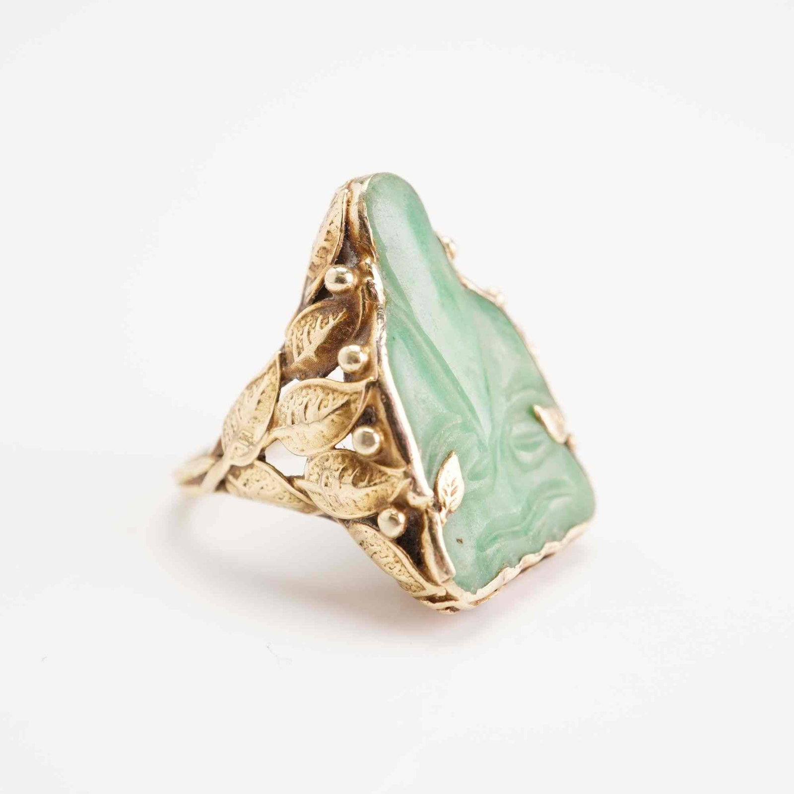 14 Karat Yellow Gold and Carved Jade Cocktail Ring: A mid-century 14 karat yellow gold cocktail ring featuring a prominent carved green jade centerpiece. The stone is carved in high relief, exhibiting a mottled light-to-medium green color. The jade is 