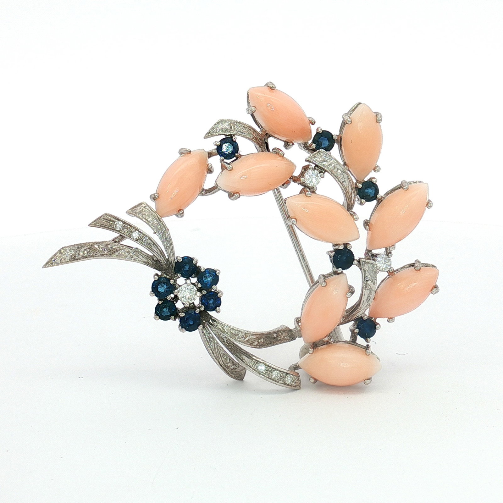 18K Gold Angel Skin Coral, Diamond, and Sapphire Brooch: A brooch crafted from 18K white gold featuring a sapphire flower with a diamond center, surrounded by ribbon-like motifs. The design includes marquise-cut angel skin coral shaped like leaves, diamond-