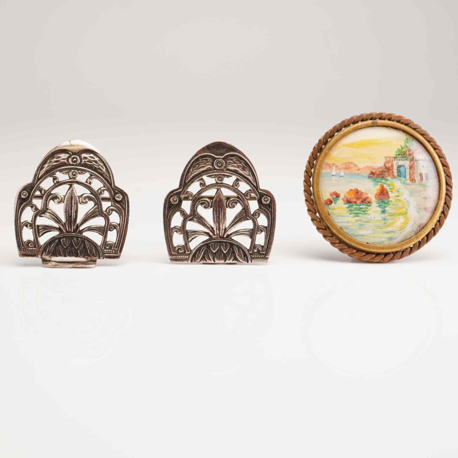 3pc Art Deco Style Dress Clips and a Hand-Painted Miniature Brooch: This lot comprises three vintage jewelry items crafted from base metals. The first component is a pair of matching dress clips in an Art Deco style. These clips feature a symmetrical openwork design w