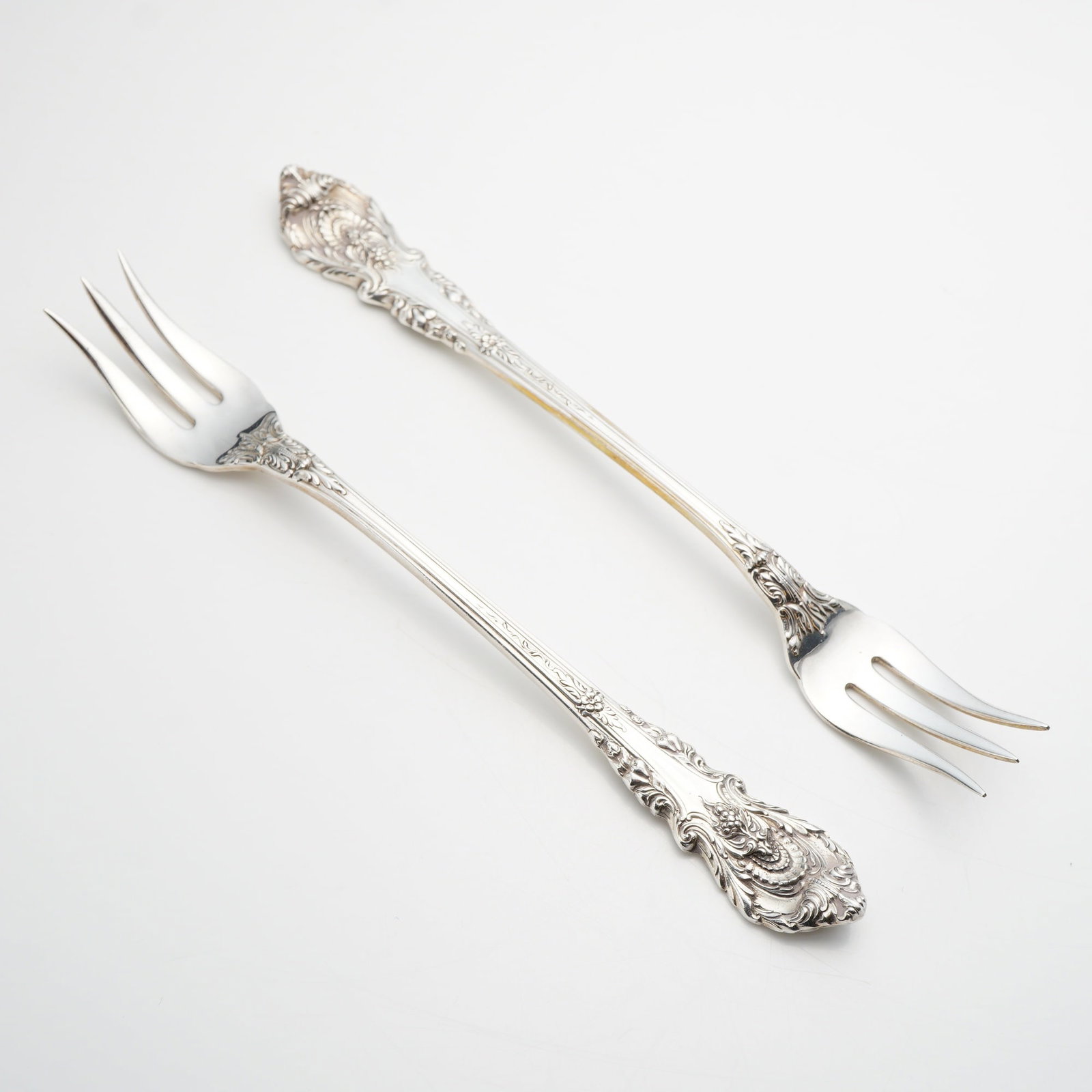 A Pair of Wallace Sterling Silver Cocktail Forks in the Sir Christopher Pattern: A pair of sterling silver cocktail or seafood forks produced by Wallace Silversmiths in the Sir Christopher pattern. Introduced in 1936, this pattern features a highly ornate, three-dimensional design