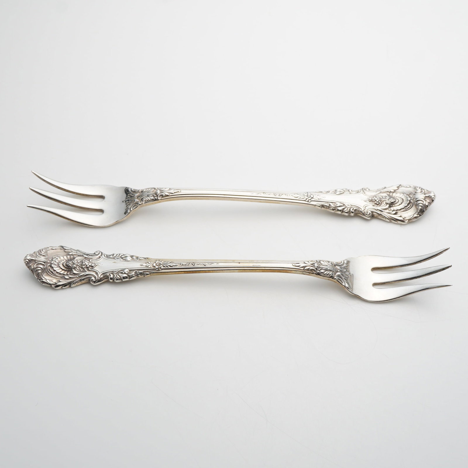 A Pair of Wallace Sterling Silver Cocktail Forks in the Sir Christopher Pattern: A pair of solid sterling silver cocktail or seafood forks produced by Wallace Silversmiths. The forks feature the Sir Christopher pattern, characterized by its ornate Baroque-inspired design with heav