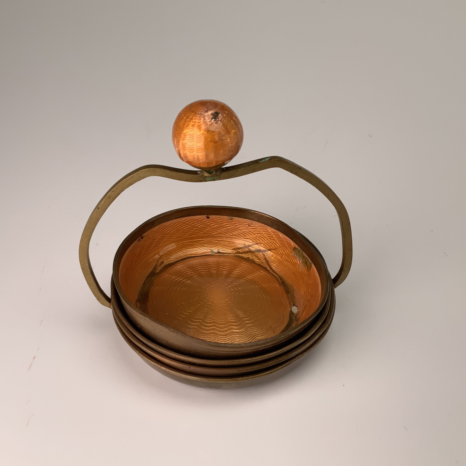 A Set of Four German Orange Guilloche Enamel and Brass Nesting Dishes: This set of four nesting dishes is constructed from brass with translucent orange guilloche enamel. The collection includes one dish with a fixed arched brass handle surmounted by a decorative sphere 