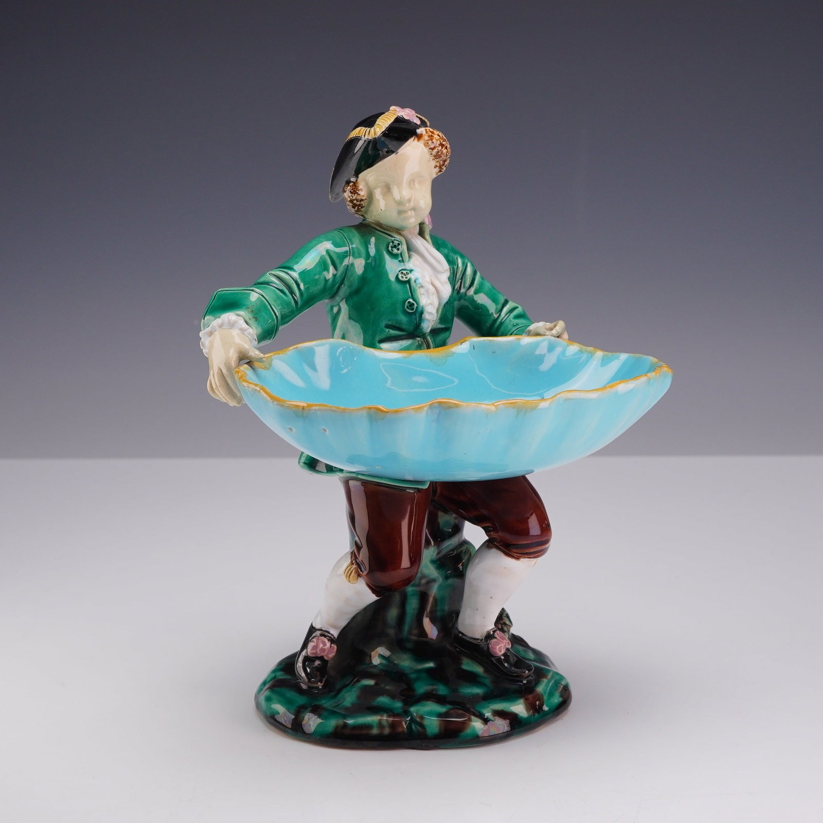 English Majolica Hogarth Boy Figurine: This majolica figurine, modeled as a Hogarth Boy, depicts a young man in 18th-century attire supporting a large scalloped shell-form bowl. The figure is dressed in a green frock coat with mottled glaz