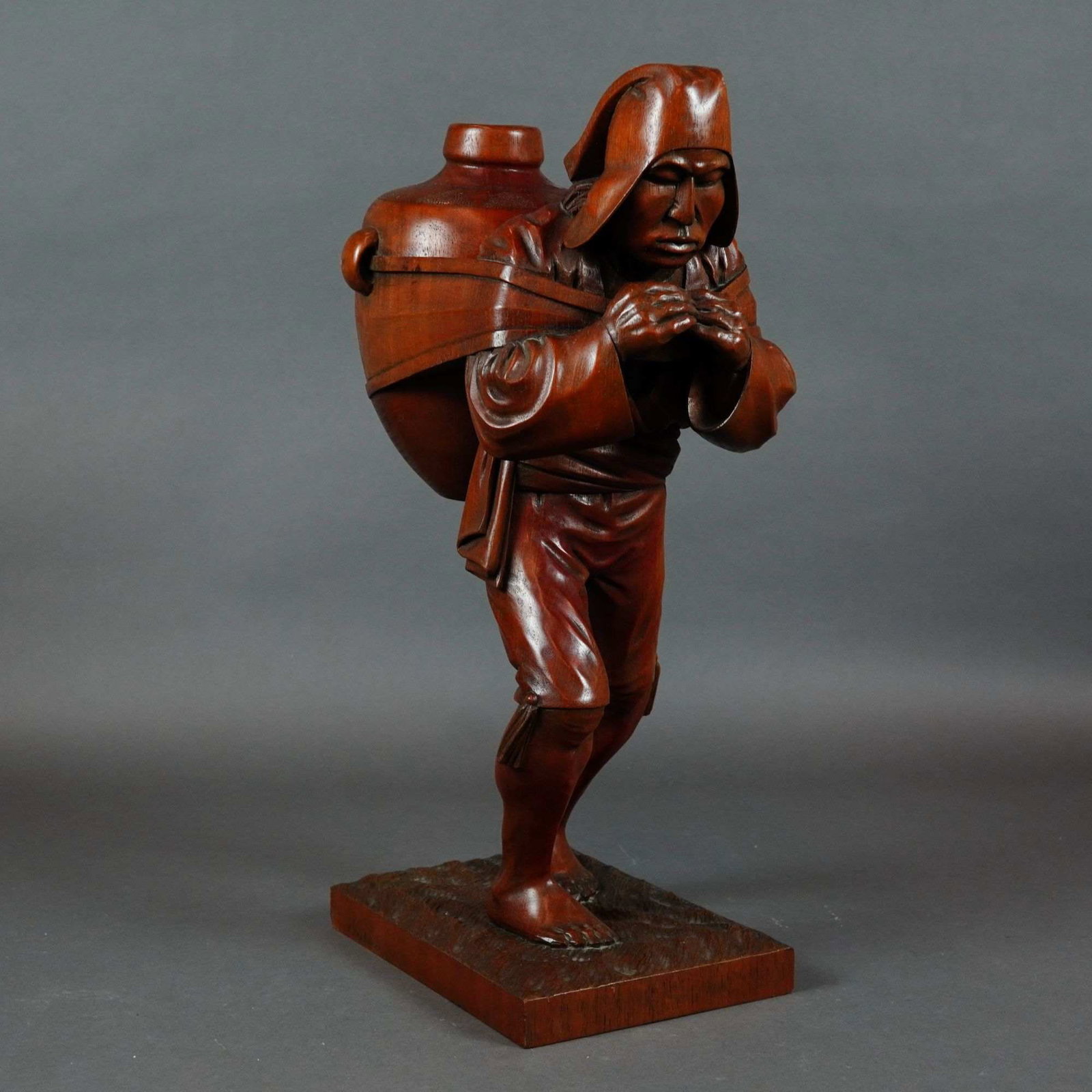 E. Pacheco Hand-Carved Wood Sculpture of a Water Carrier: This hand-carved wooden sculpture by E. Pacheco depicts a laborer or water carrier in a dynamic, forward-leaning stance. The figure carries a large, heavy vessel on his back, secured by a wide strap a