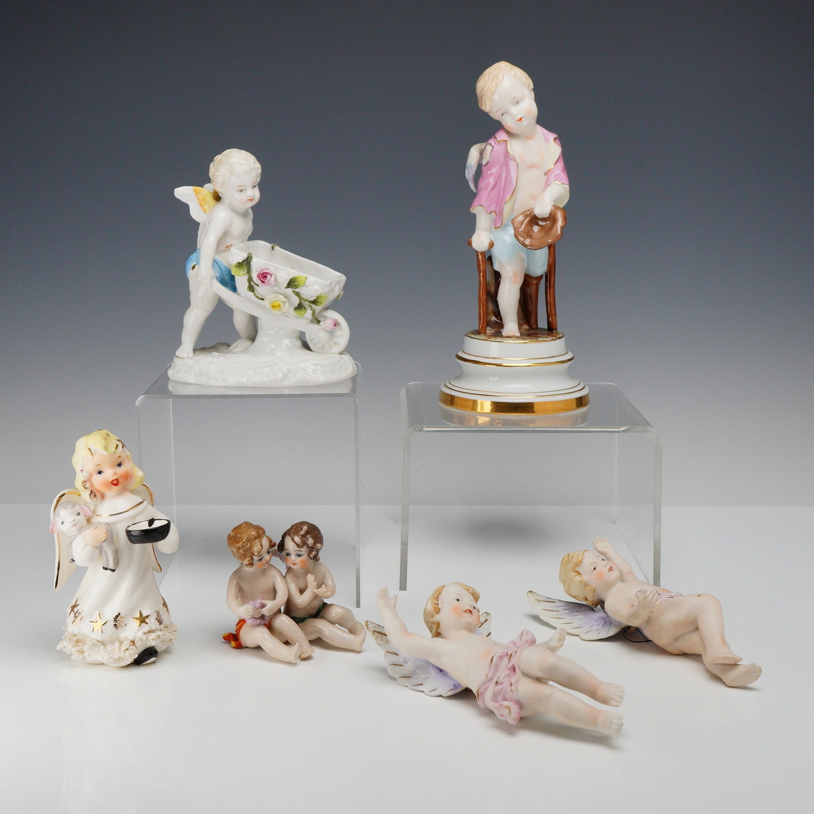 Mixed Lot of 6 Porcelain Angel and Cherub Figurines: This collection consists of six glazed and hand-painted porcelain figurines depicting various angels and cherubs. The lot includes a standing winged cherub on a circular pedestal with gold-gilt bands,