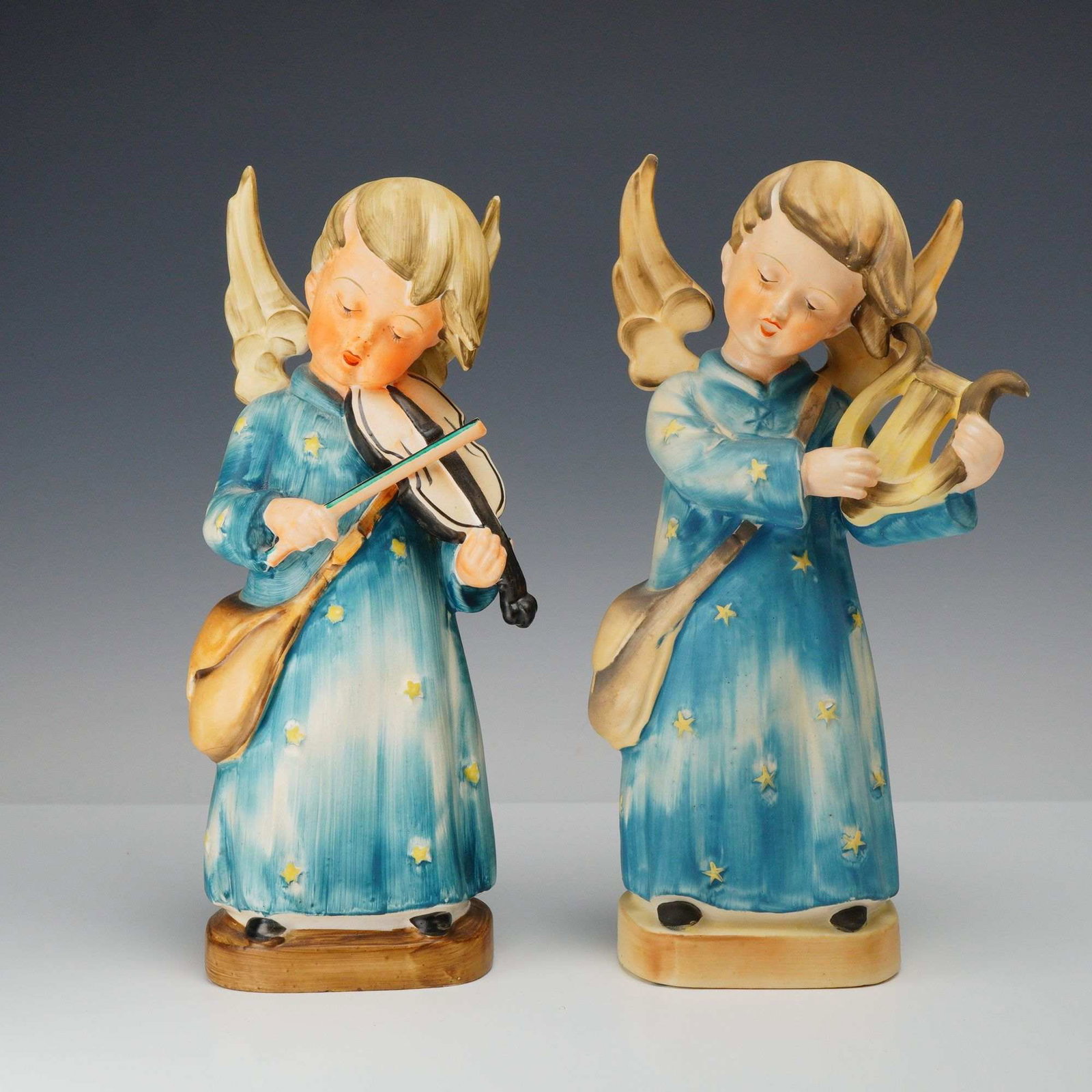 Lot of 2 Hummel-Style Ceramic Angel Figurines Holding Musical Instruments: This lot consists of two polychrome glazed ceramic figurines of angels, executed in a style reminiscent of mid-century European collectibles. Each figure features a child-like angel with blonde hair a