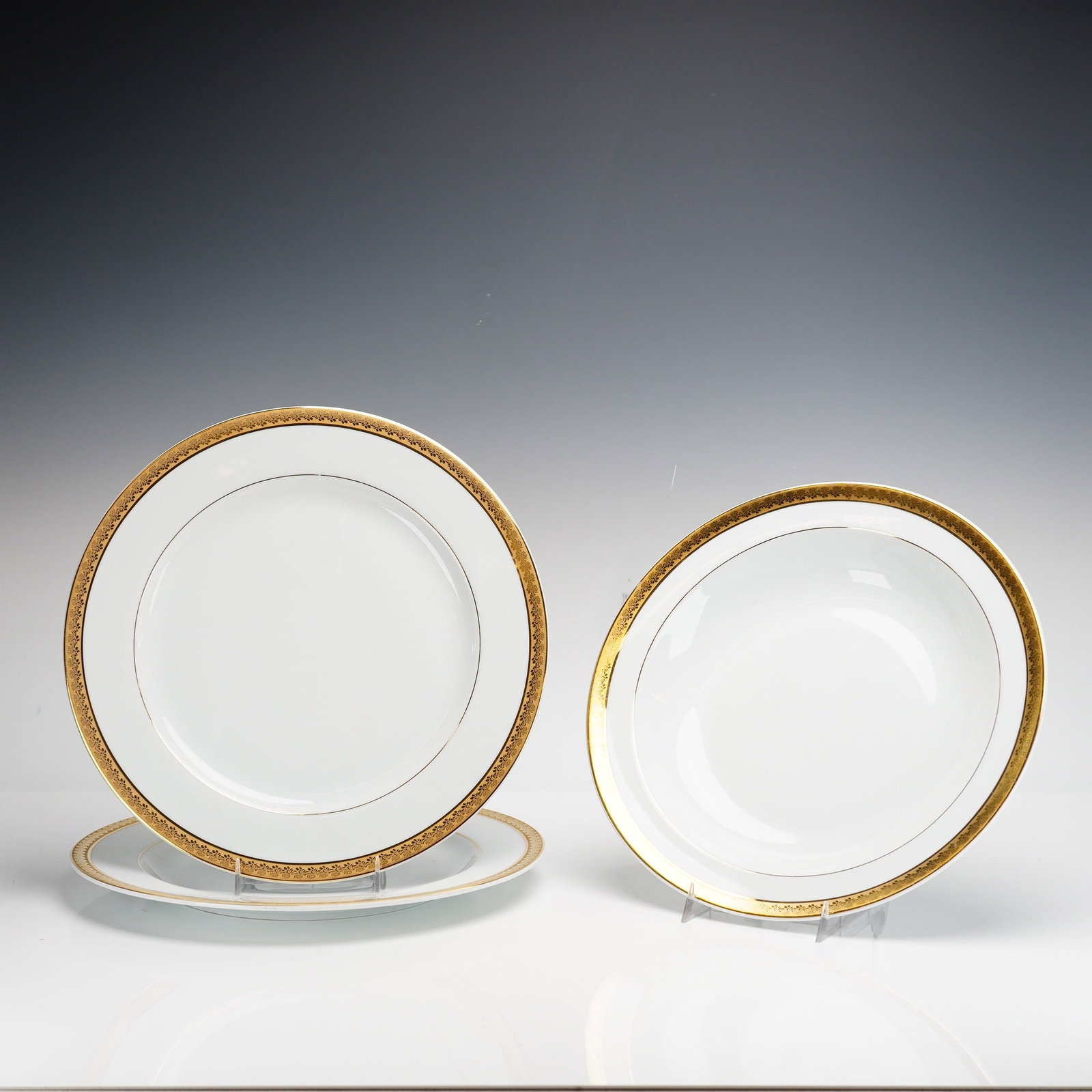 Set of Three Bernardaud Limoges Porcelain Chargers and Serving Bowl: The set comprises two circular charger plates and one rimmed serving bowl. Each piece is constructed from white porcelain with a wide, ornate gilded border. The decoration consists of an intricate, re