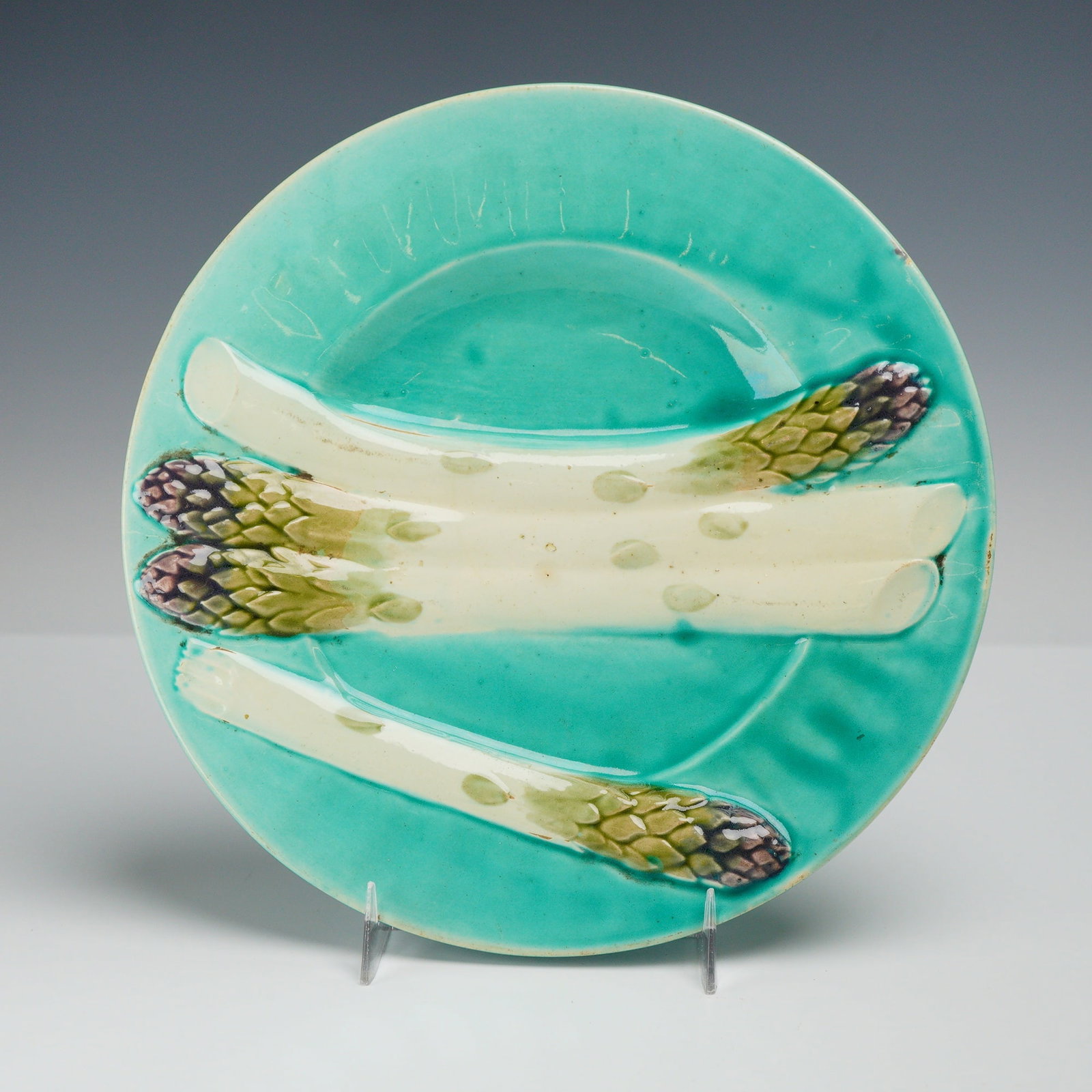 French Luneville Majolica Asparagus Plate: This glazed earthenware plate features a vibrant turquoise ground decorated with four relief-molded asparagus spears. The spears are finished in naturalistic shades of cream, green, and purple. The