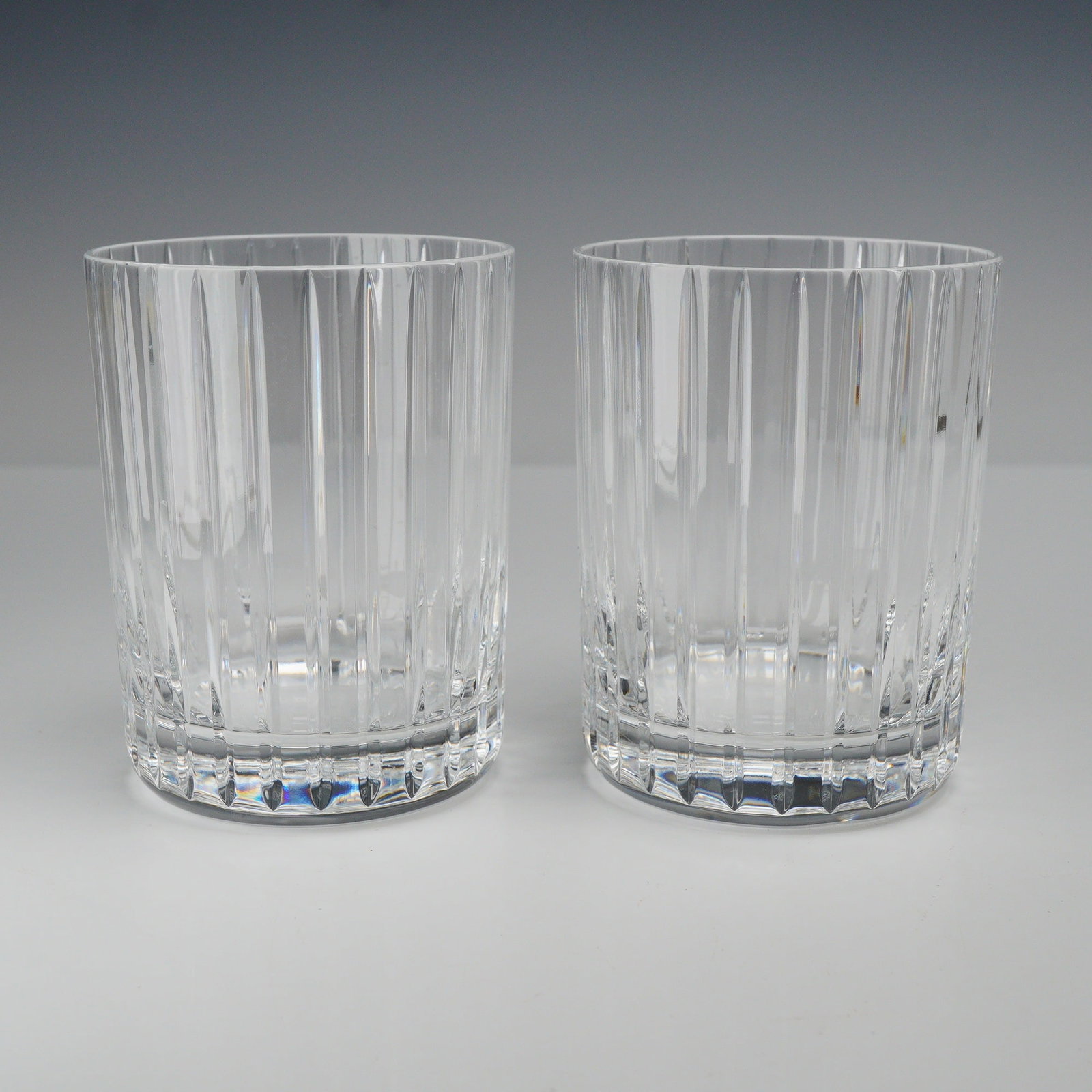 2pc Baccarat Cut Crystal Harmonie Tumbler Whiskey Glasses: This lot consists of two Baccarat cut crystal tumblers in the Harmonie pattern. Crafted from clear lead crystal, each glass is decorated with a series of parallel vertical flutes, characterized by dee