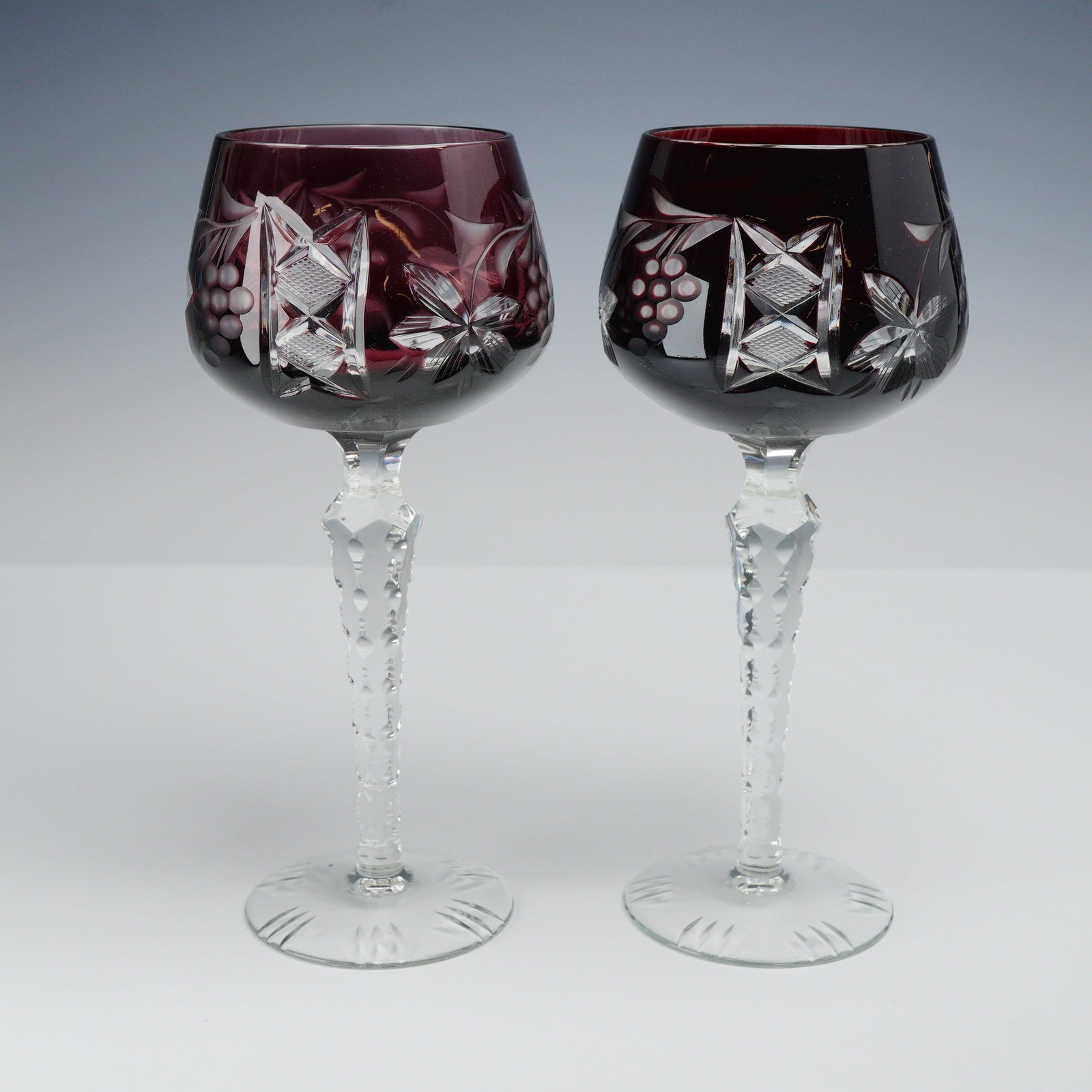 2pc Cut-to-Clear Amethyst Crystal Hock Wine Glasses: The bowls are decorated with a hand-cut pattern including grape clusters and stylized vine leaves interspersed with geometric star motifs containing cross-hatched and diamond cuts. The glasses