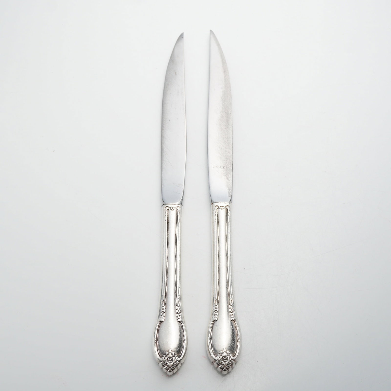 Two Sterling Silver Handled Knives with Floral and Scroll Decoration: This lot of 2 sterling silver knives consists of steak knives featuring ornate, hollow-form handles and stainless steel blades. The decoration is rendered in a Rococo-revival style, characterized by
