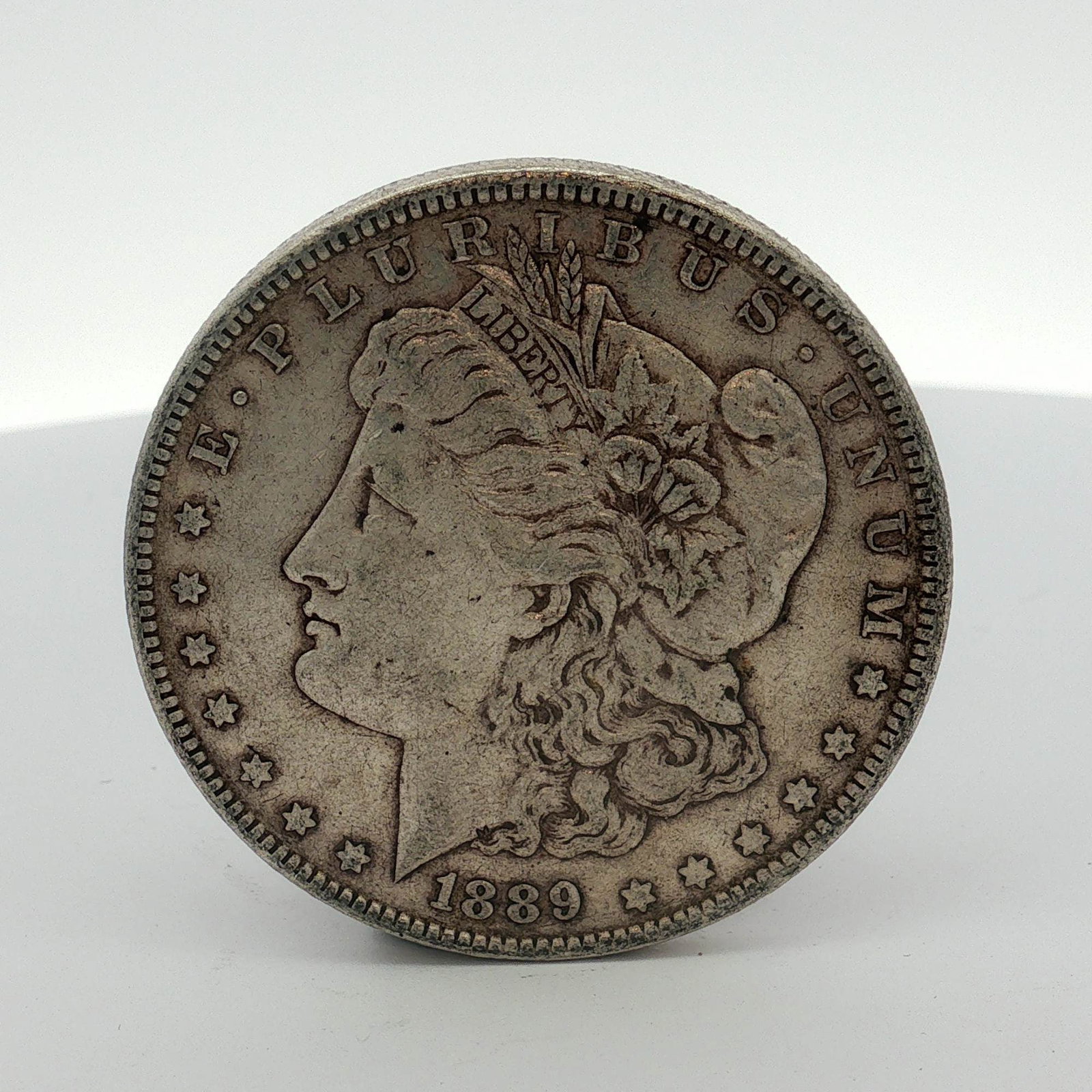 1889 US Morgan Silver Dollar Coin Circulated: This US 1889-O Morgan dollar is a 90% silver coin struck at the Philadelphia Mint. The specimen features the Morgan dollar design type. The obverse is inscribed E PLURIBUS UNUM and dated 1889. The rev