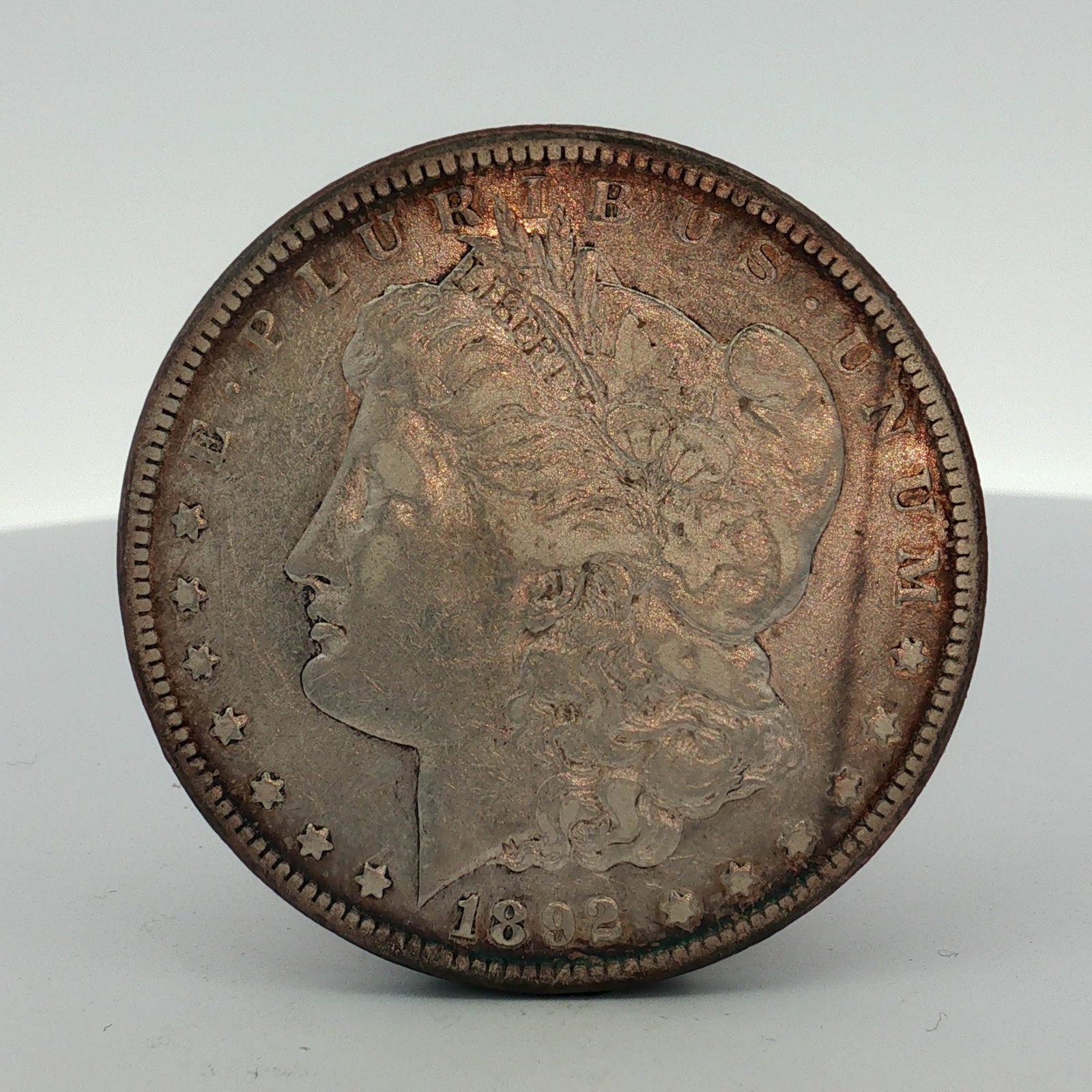 1892-CC US Morgan Silver Dollar: This 1892-CC US Morgan Carson City dollar was struck at the Carson City Mint and is composed of .900 fine silver. The obverse presents the Morgan Liberty head design with the date 1892 and thirteen st