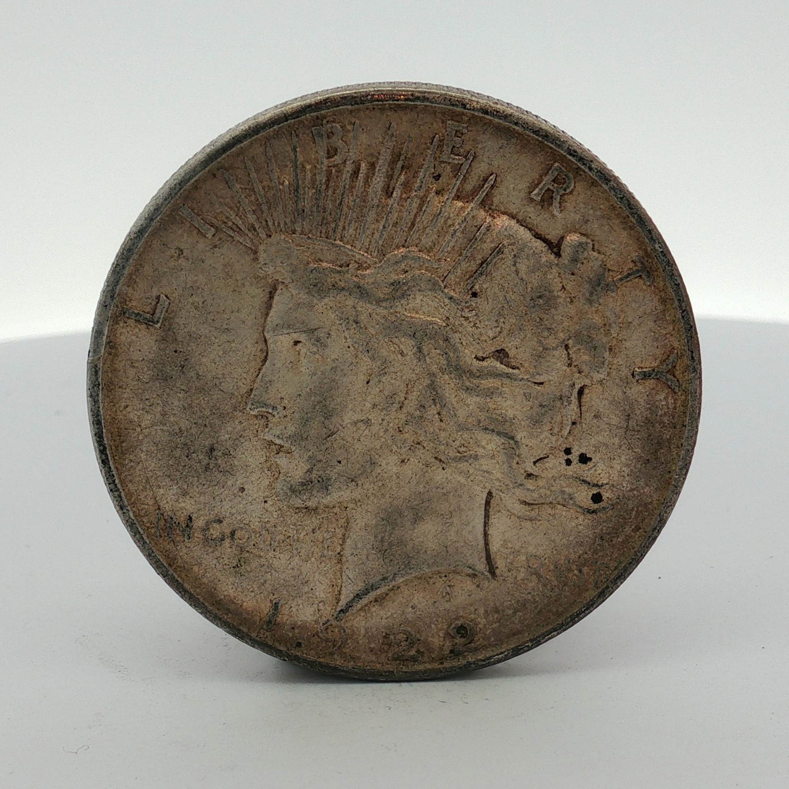 1922-D US One Dollar Silver Peace Dollar, Circulated: This US Peace silver dollar was struck at the Denver Mint in 1922. The coin is composed of silver and features a reeded edge. The obverse displays the Liberty head design with inscriptions LIBERTY, IN