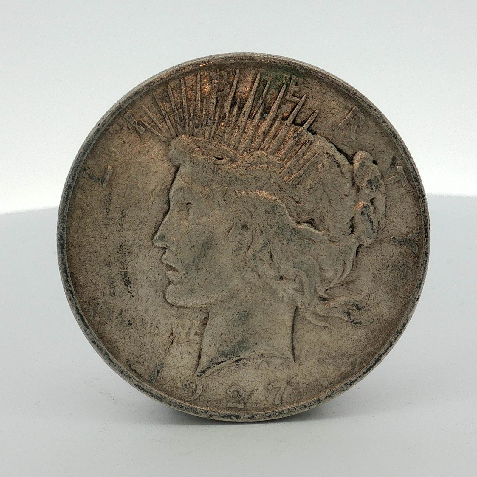 1927-D US One Dollar Peace Silver Dollar, Circulated: This 1927 US Peace silver dollar was struck at the Denver Mint. The coin is composed of 90% silver. The obverse features the Liberty design and the reverse features the Peace eagle design. Inscription