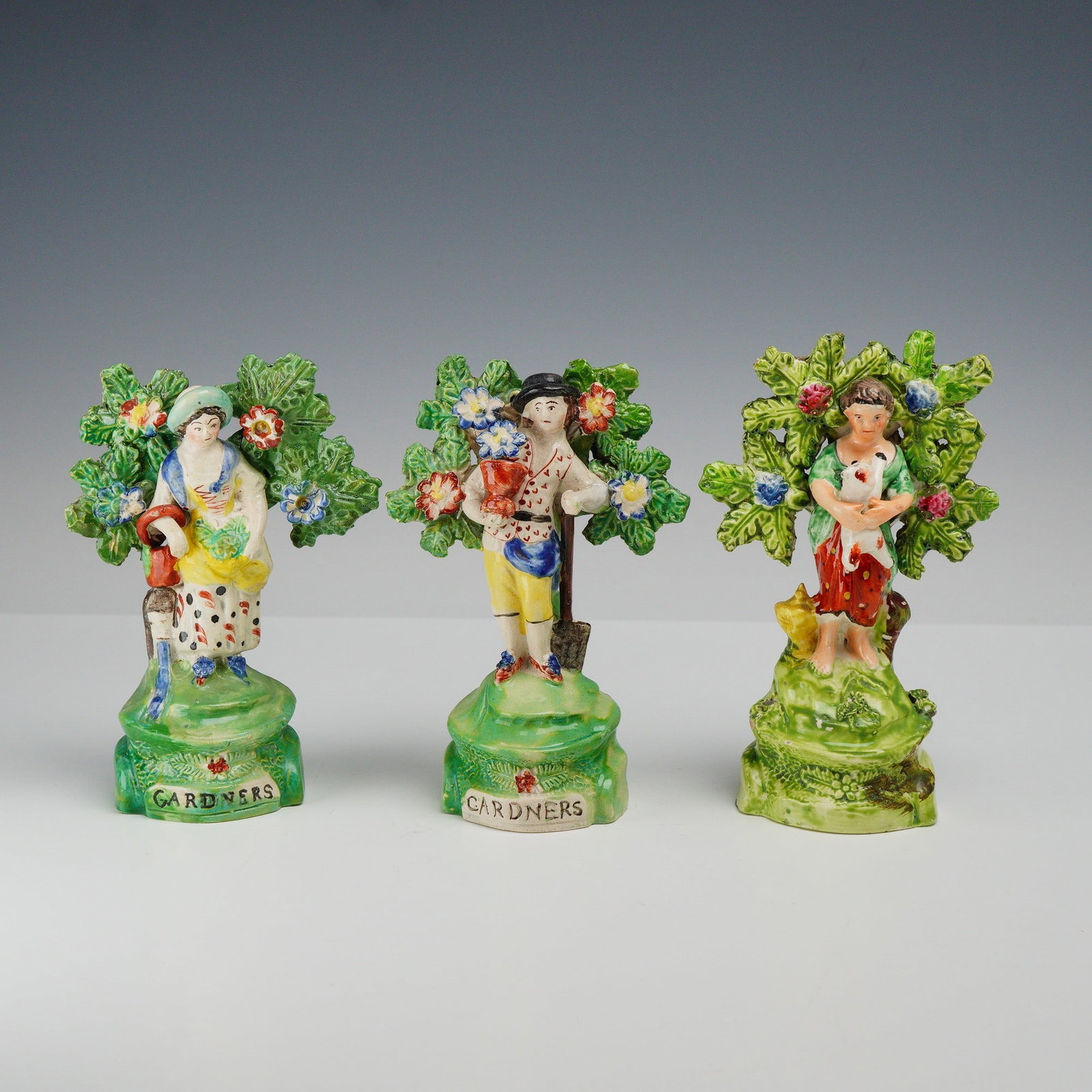 3pc Staffordshire Pottery Figurines, Gardeners & Child w/ Lamb: The collection includes a pair of figures titled GARDNERS and a figure of a child with a lamb. The female gardener is depicted standing before a flowering bocage, wearing a skirt with black spots, a w