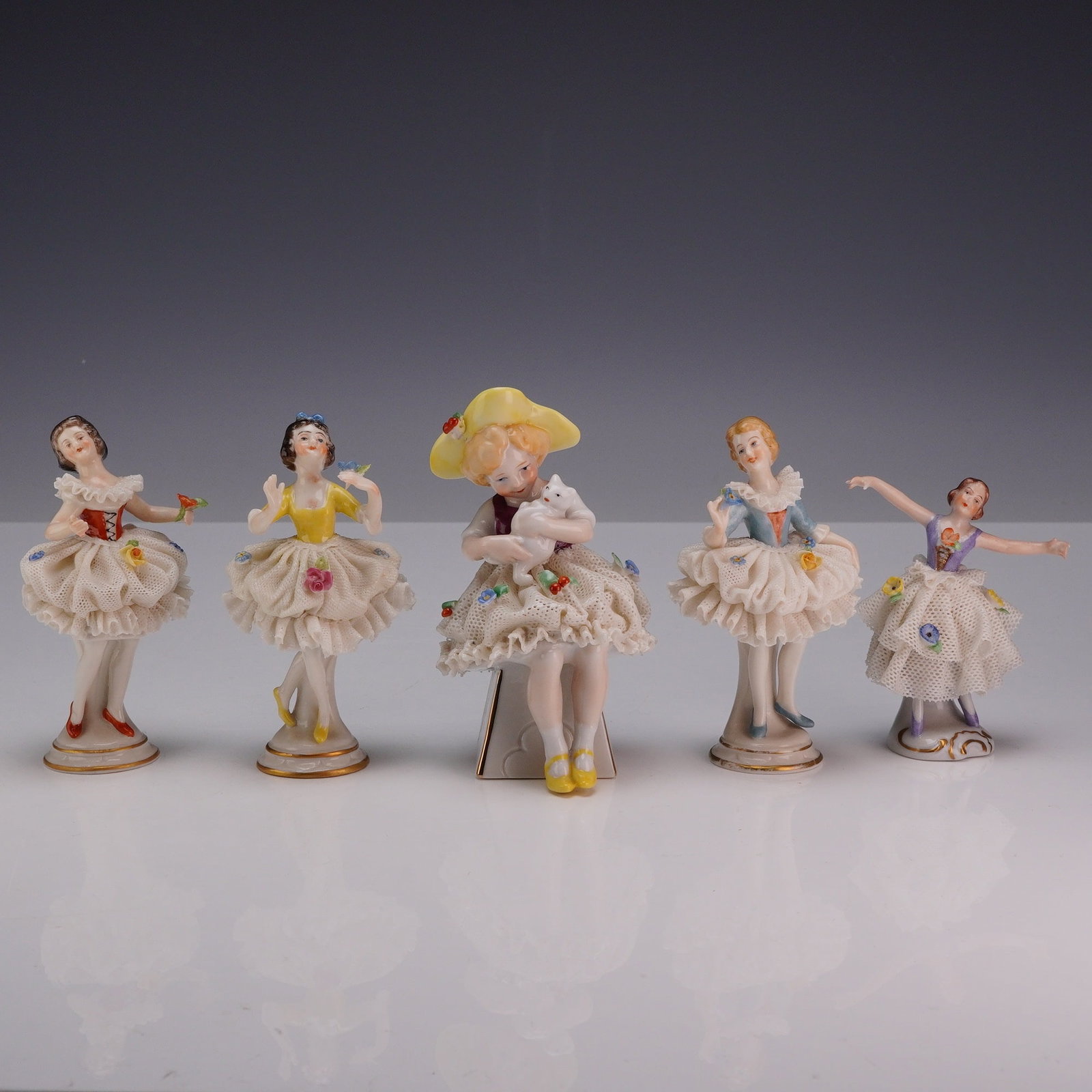 5pc Germany Porcelain Little Ladies Figurines: Collection of hand-decorated German porcelain figurines depicting elegant young ladies in flowing ruffled skirts accented with applied floral relief decoration. The grouping includes three figures by 