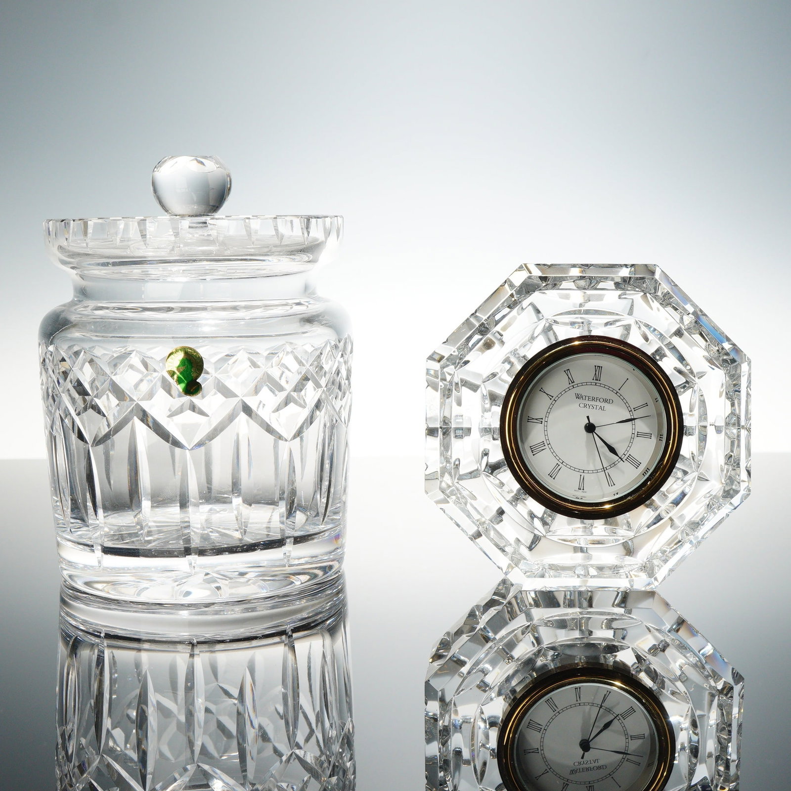 Two Waterford Crystal Pieces including a Lismore Pattern Biscuit Barrel and an Octagonal Desk Clock: This lot contains two pieces of hand-cut lead crystal manufactured by Waterford. The first item is a biscuit barrel, decorated in the Lismore pattern. The design features deep vertical wedge cuts on t