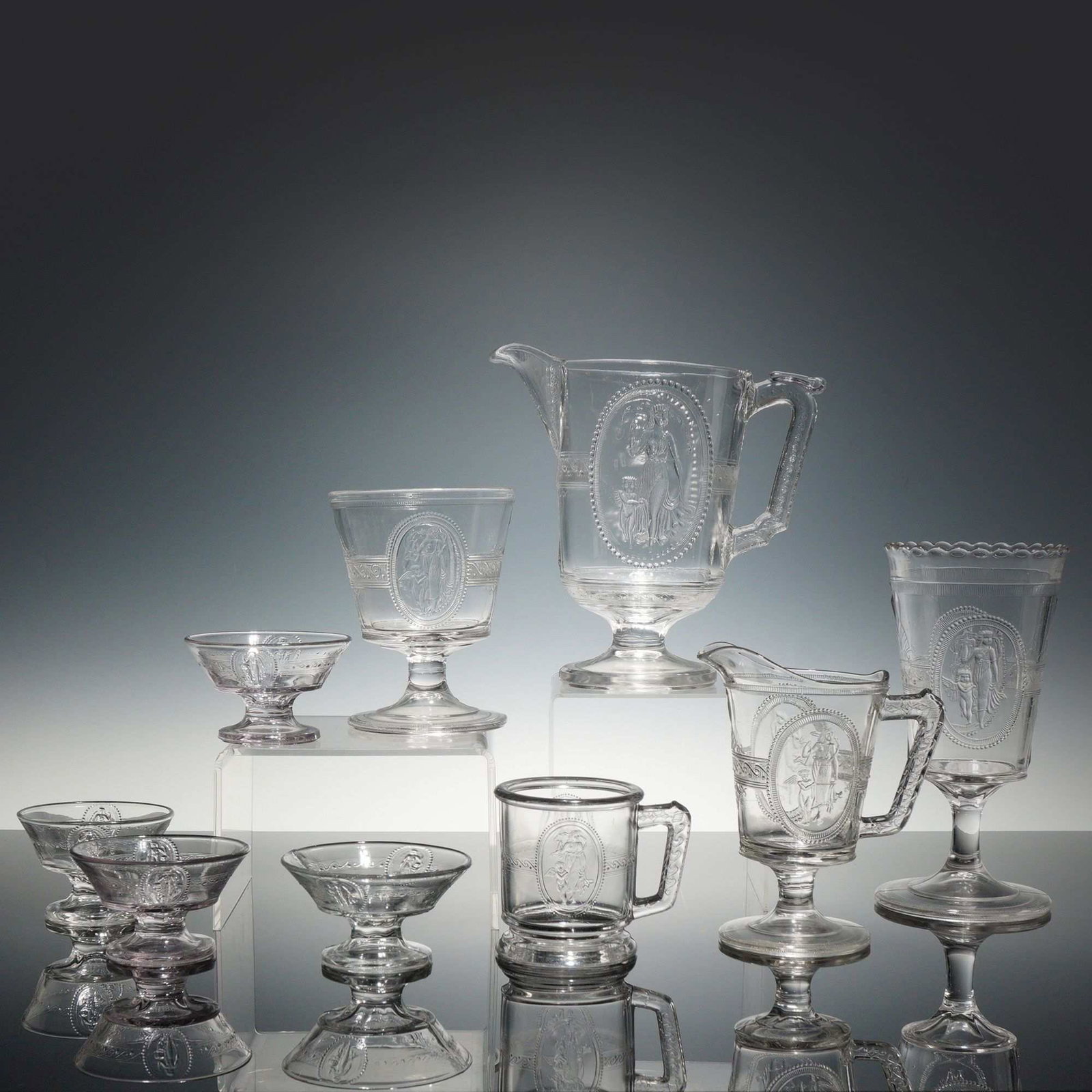 Set of 9 Richards and Hartley Pressed Glass Pitchers and Cups, Cupid and Venus Pattern: This is a set of 9 pieces of Early American Pressed Glass (EAPG) in the Cupid and Venus pattern, also known as Guardian Angel, manufactured by the Richards and Hartley Flint Glass Company of Pittsburg
