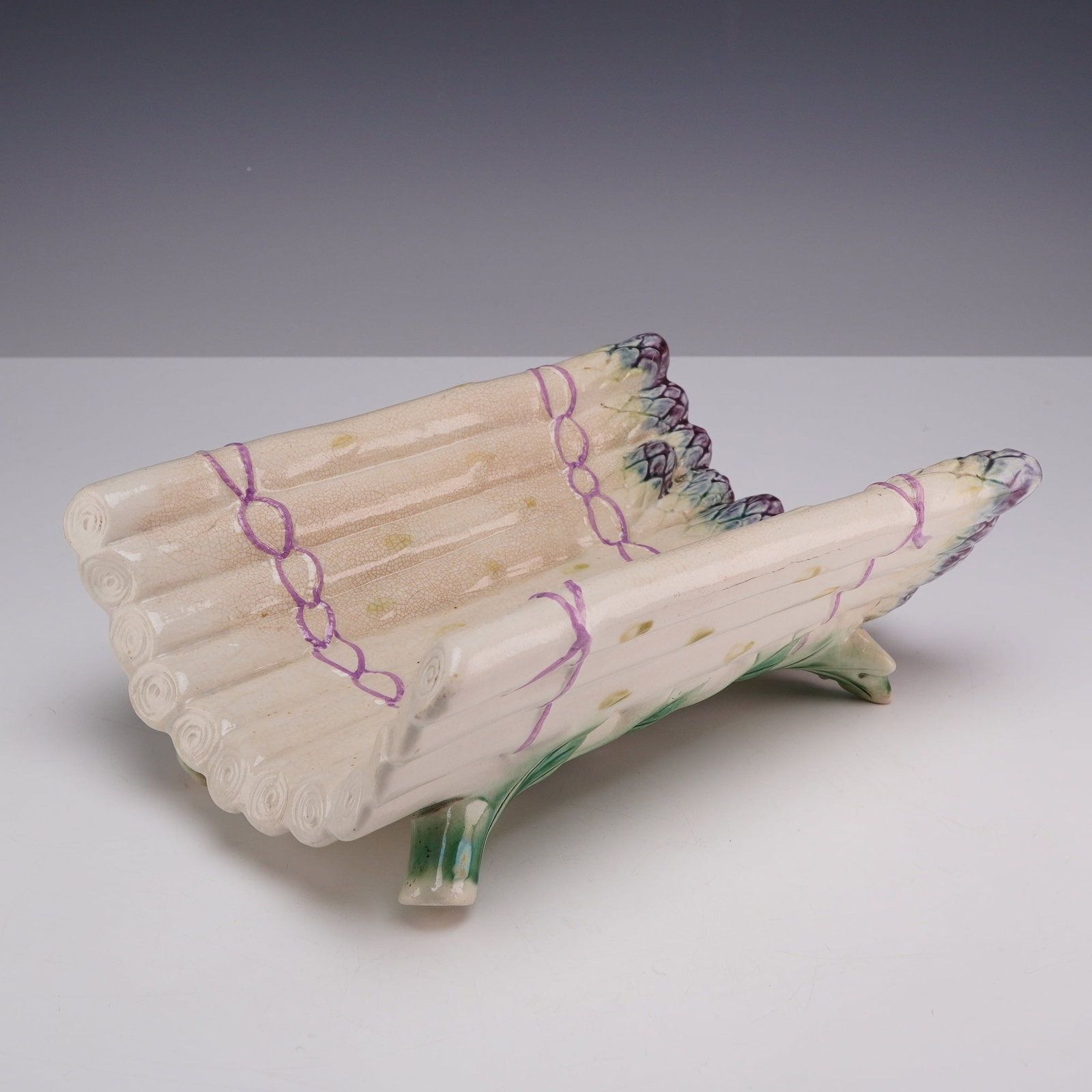 French Style Majolica Asparagus Tray: This majolica-style asparagus tray is a piece of glazed earthenware modeled as a curved cradle. The design simulates a bundle of white asparagus stalks tied together with two rows of purple ribbons.