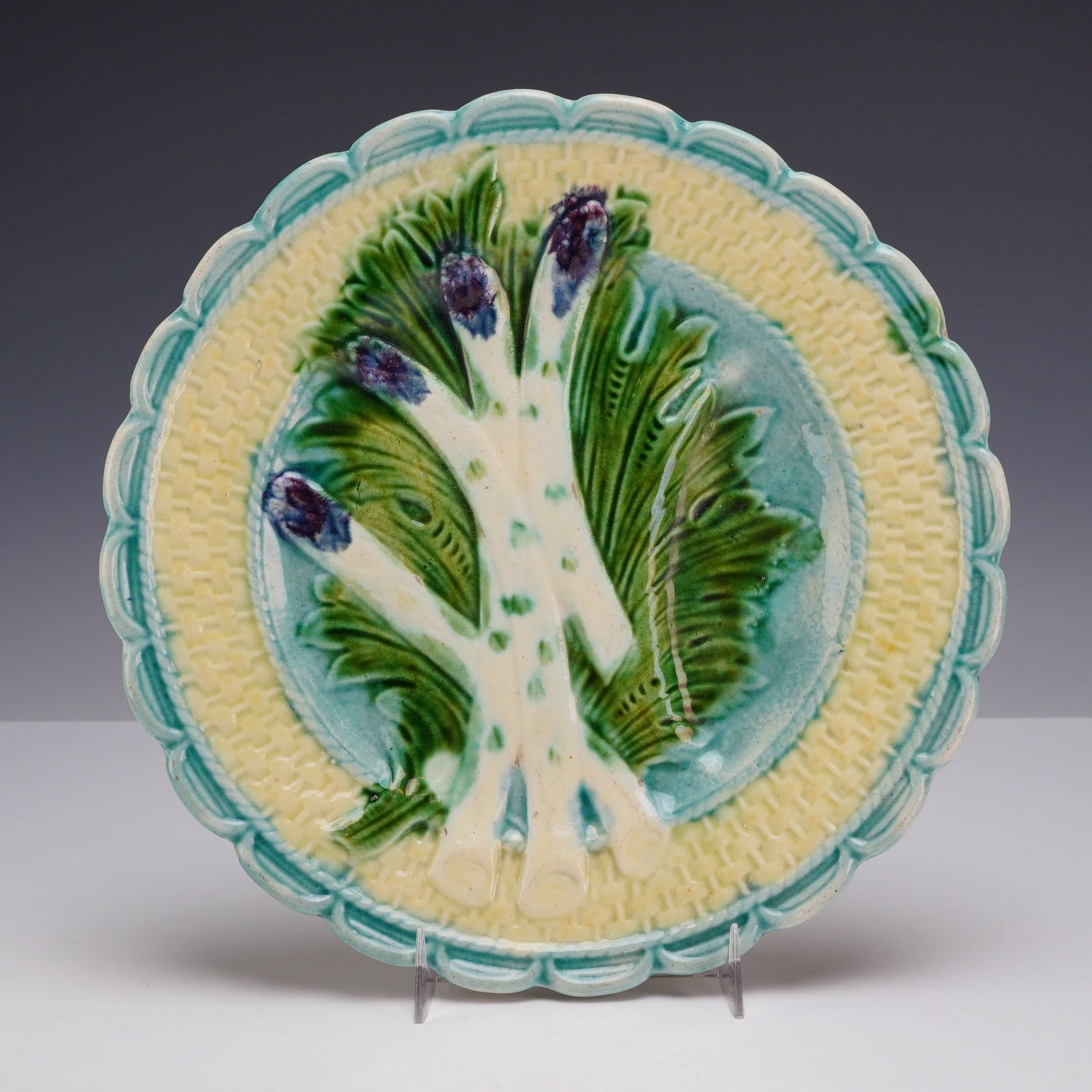 French Majolica Asparagus Plate: A circular lead-glazed earthenware majolica plate featuring a relief-molded design. The central motif consists of four white asparagus stalks with purple-blue tips and green flecks, set against a