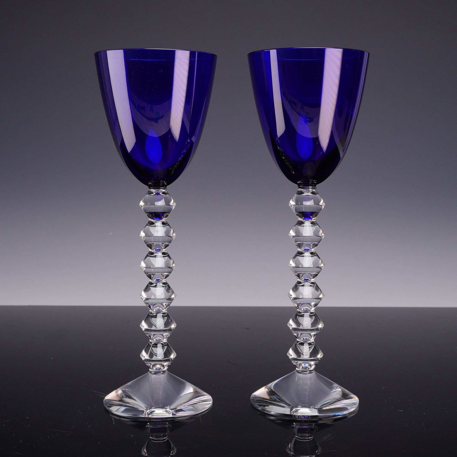 2pc Baccarat Vega Cobalt Blue Stemmed Wine Glasses: A pair of Baccarat crystal wine glasses in the Vega pattern, featuring deep cobalt blue bowls and clear crystal stems. Each glass is designed with a U-shaped bowl set upon a stem composed of six stack