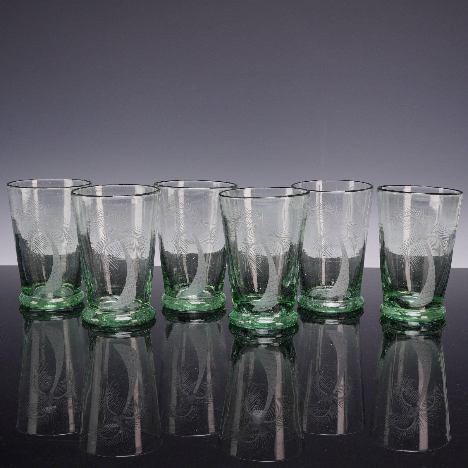 6pc Coconut Tree Design Drink Glasses in Hand-Blown Green Glass: This set of six hand-blown artisan tumblers is crafted from translucent pale green glass, featuring a distinctive etched coconut tree design on each piece. The glass material exhibits characteristic