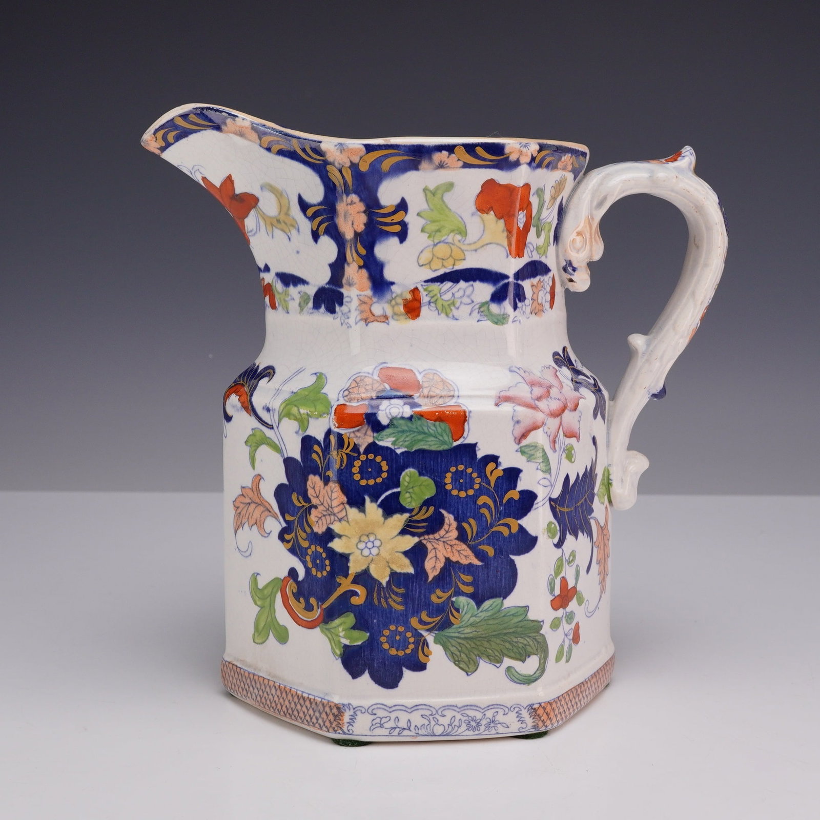 English New Stone China Ironstone Pitcher: An English New Stone China ironstone pitcher featuring a hexagonal-paneled body that tapers to a waisted neck and a wide, flared spout with a scalloped rim. The vessel is equipped with a heavily