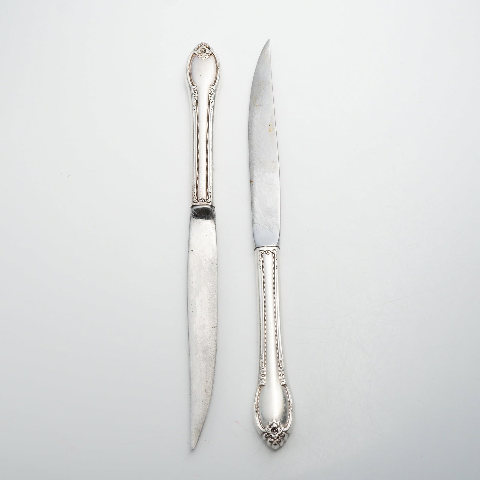 Two Sterling Silver Handled Knives with Floral and Scroll Decoration: A pair of knives featuring sterling silver handles and stainless steel blades. The handles are designed in a traditional American floral repoussé style, characterized by a central multi-petaled flowe