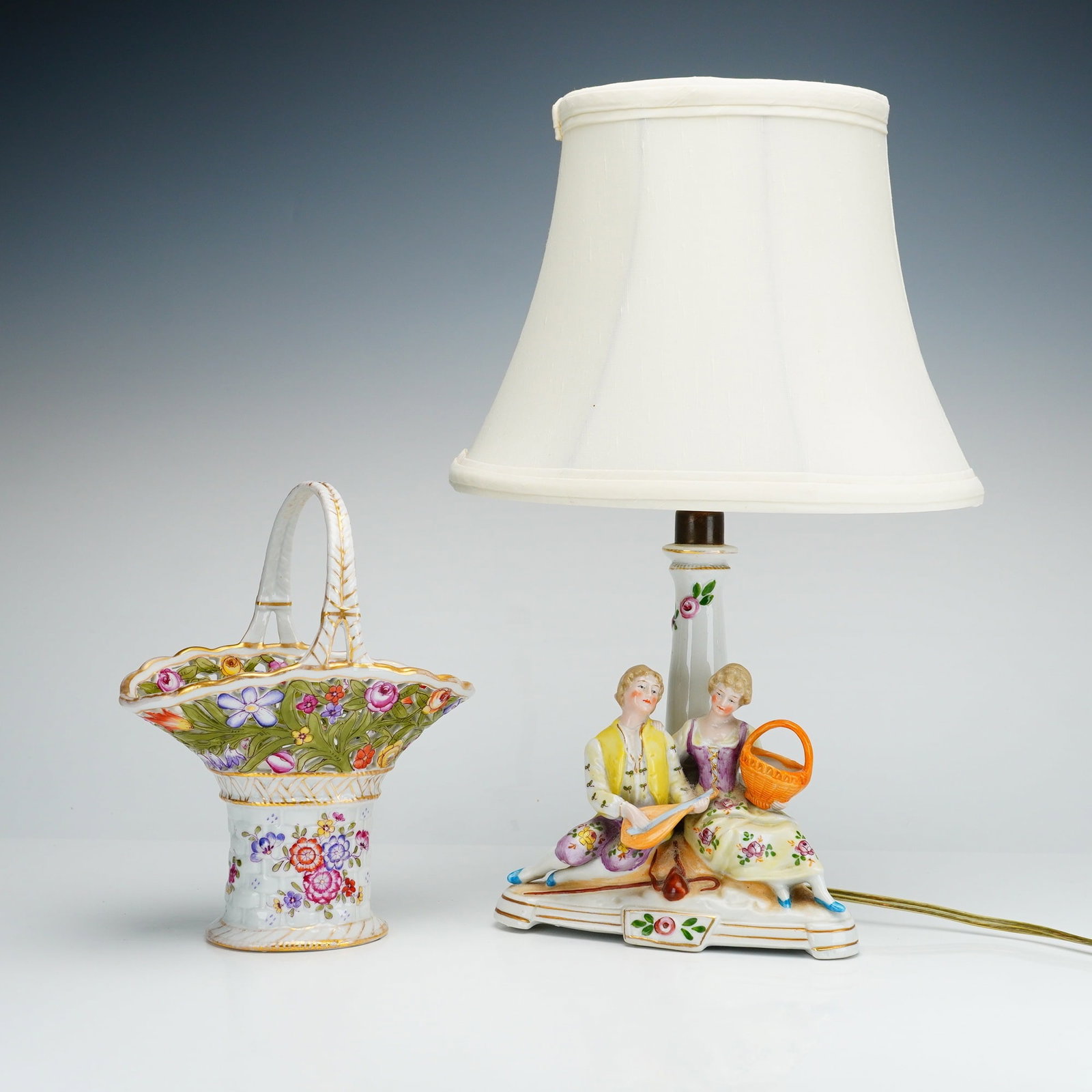 2pc German Items, Von Schierholz Basket and a Victorian Figural Lamp: This lot features a Von Schierholz porcelain basket and a Victorian porcelain figural table lamp. The hand-painted porcelain basket is designed with an openwork pierced lattice rim and a flared, scall