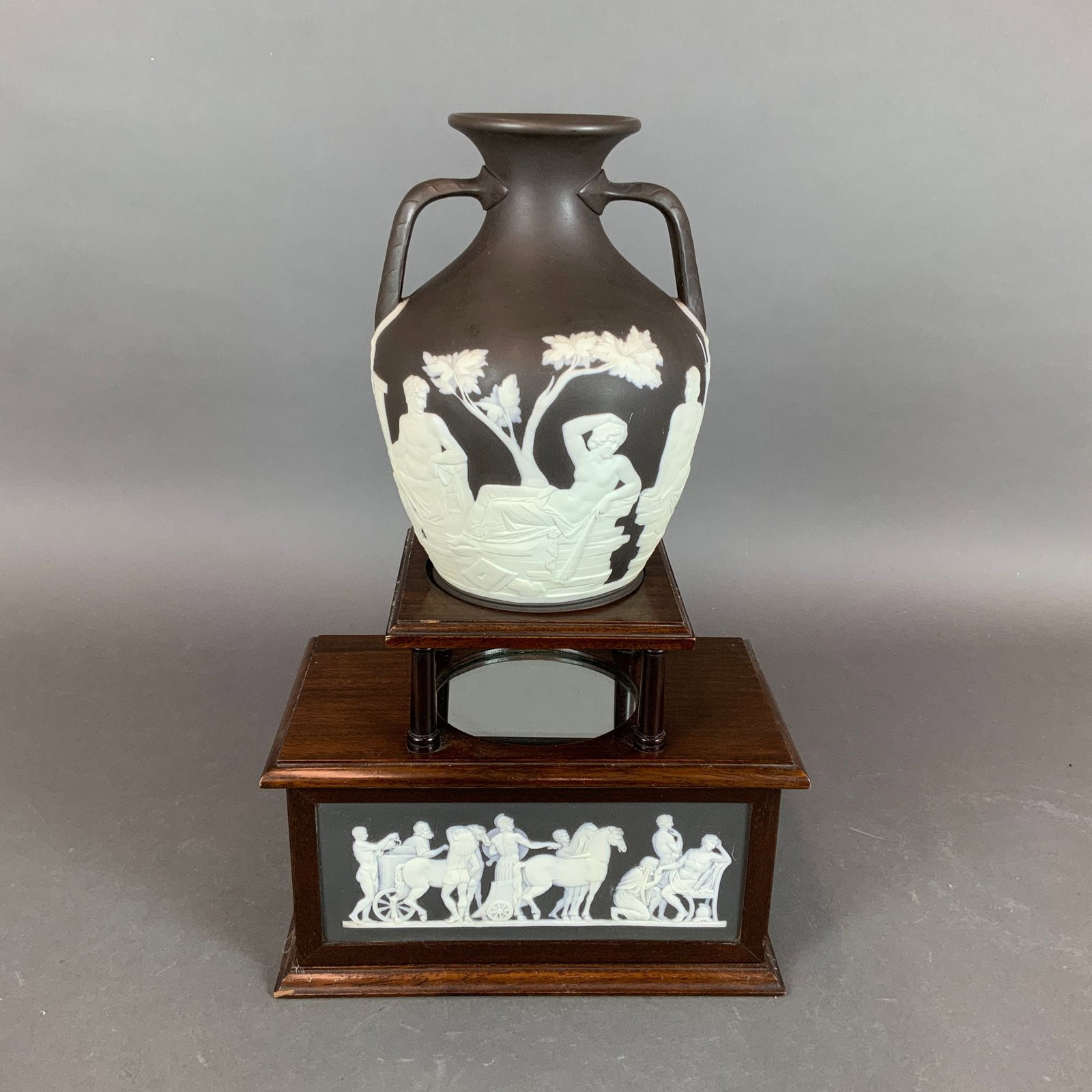 A Wedgwood Black Jasperware Portland Vase and Sarcophagus Stand: This lot consists of a Wedgwood black jasperware vase in the iconic Portland form, accompanied by a custom-designed wooden sarcophagus-style pedestal stand. The ovoid, twin-handled vase is crafted fro