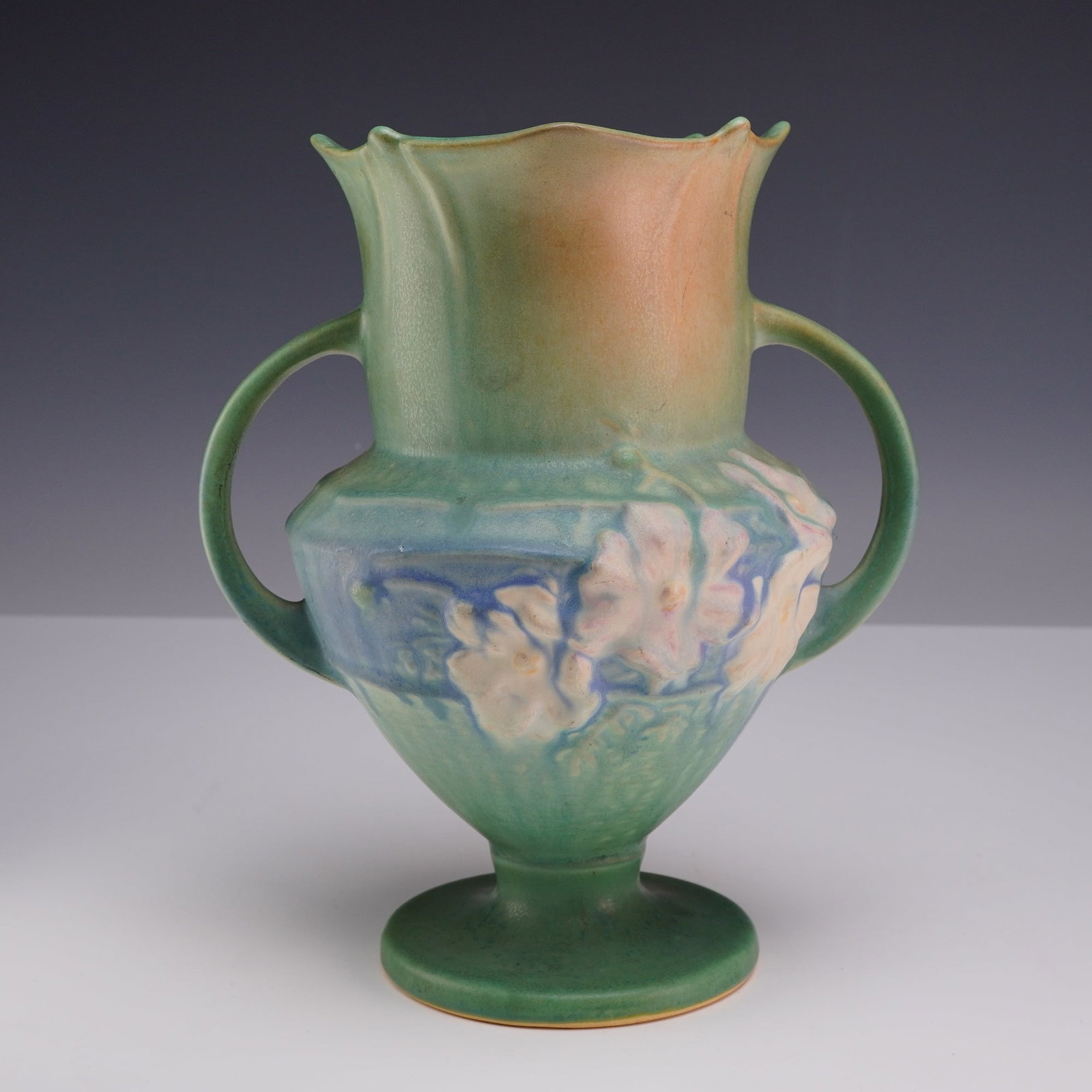 Roseville Cosmos Double Handle Art Pottery Vase: The earthenware vessel features an urn-style form with a flared, scalloped rim and a pedestal base. The body is decorated with molded floral sprays depicting clusters of white and pink cosmos blossoms