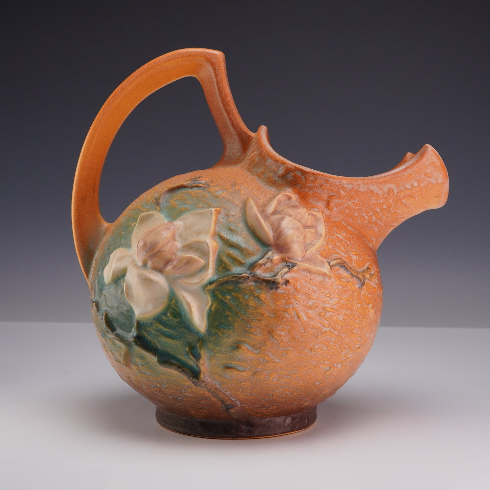 Roseville Pottery Magnolia Brown Pattern Ice Lip Pitcher Shape 1327: A Roseville Pottery earthenware pitcher in the Magnolia pattern, shape number 1327, introduced circa 1943. The vessel features a bulbous, textured body with a matte glaze in a variegated orange and