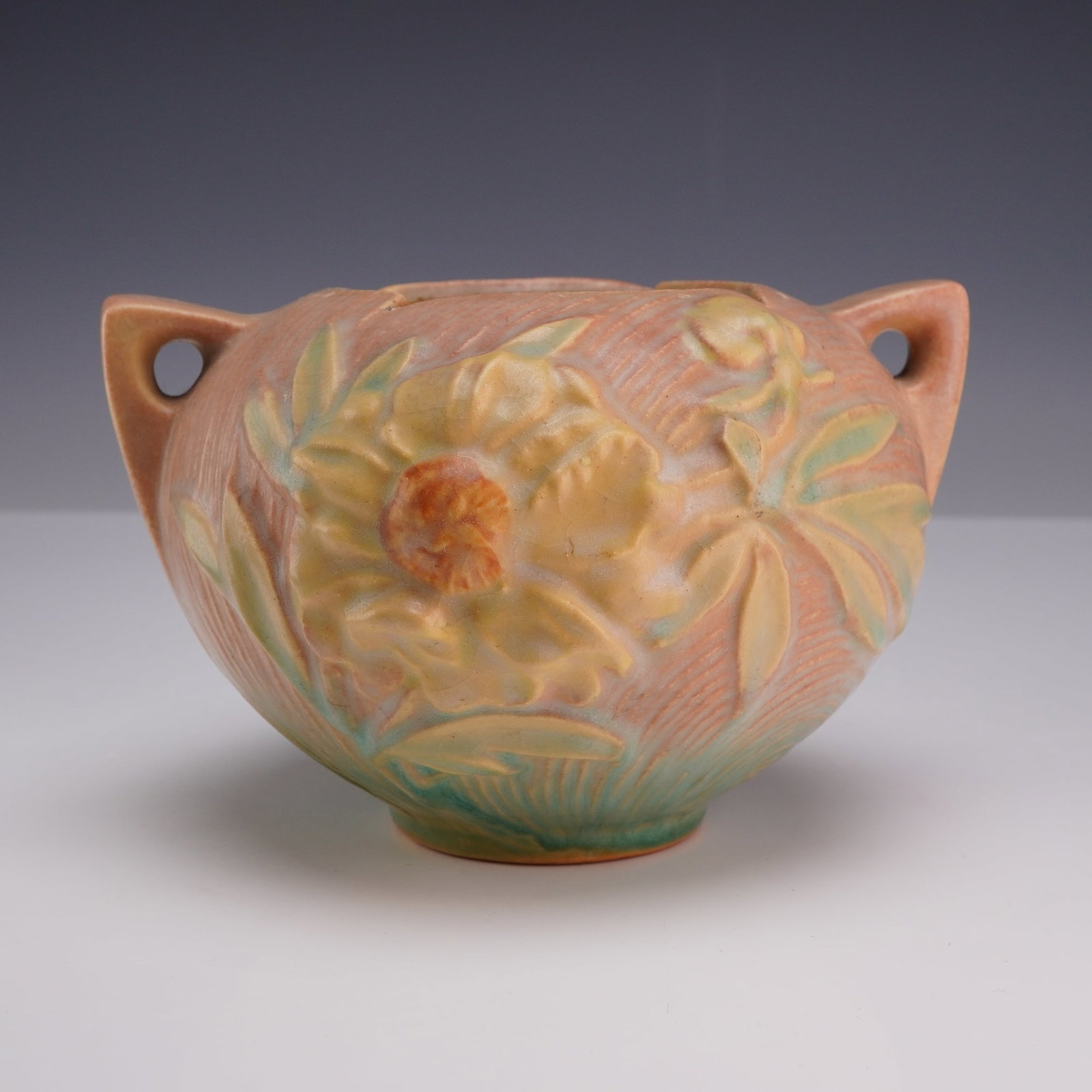 Roseville Pottery Peony Pattern Two-Handled Coral Pink and Green Rose Bowl 427-4: A glazed earthenware bowl vase produced by Roseville Pottery in the Peony pattern, which was introduced in 1942. The vessel features a bulbous form with two integral, angular handles located at the ri