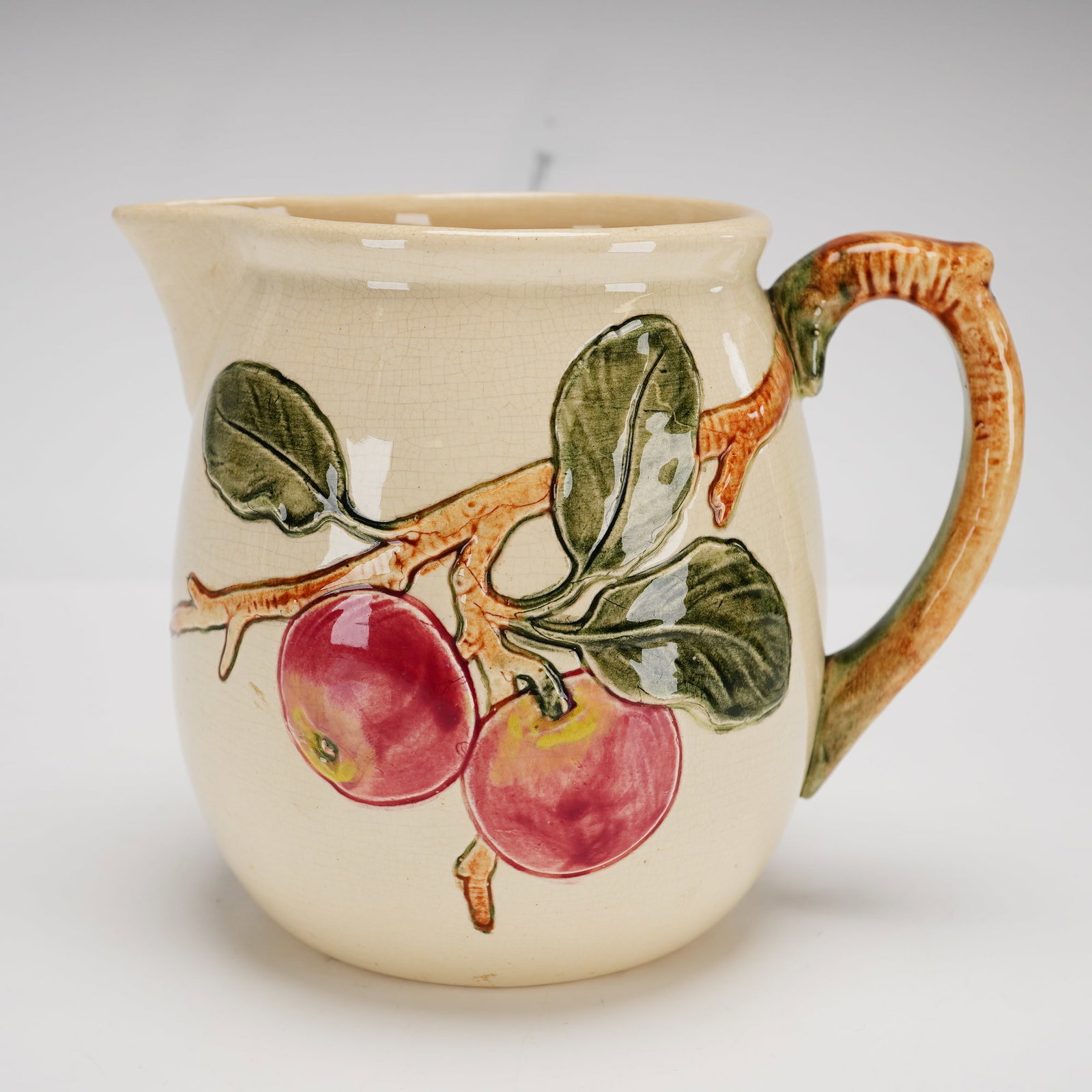 Weller Zona Apple Pattern Earthenware Pitcher: This earthenware pitcher is produced in the Zona apple pattern by Weller. The vessel features a bulbous body with a pinched spout and relief-molded decoration. The hand-painted design depicts ripe red