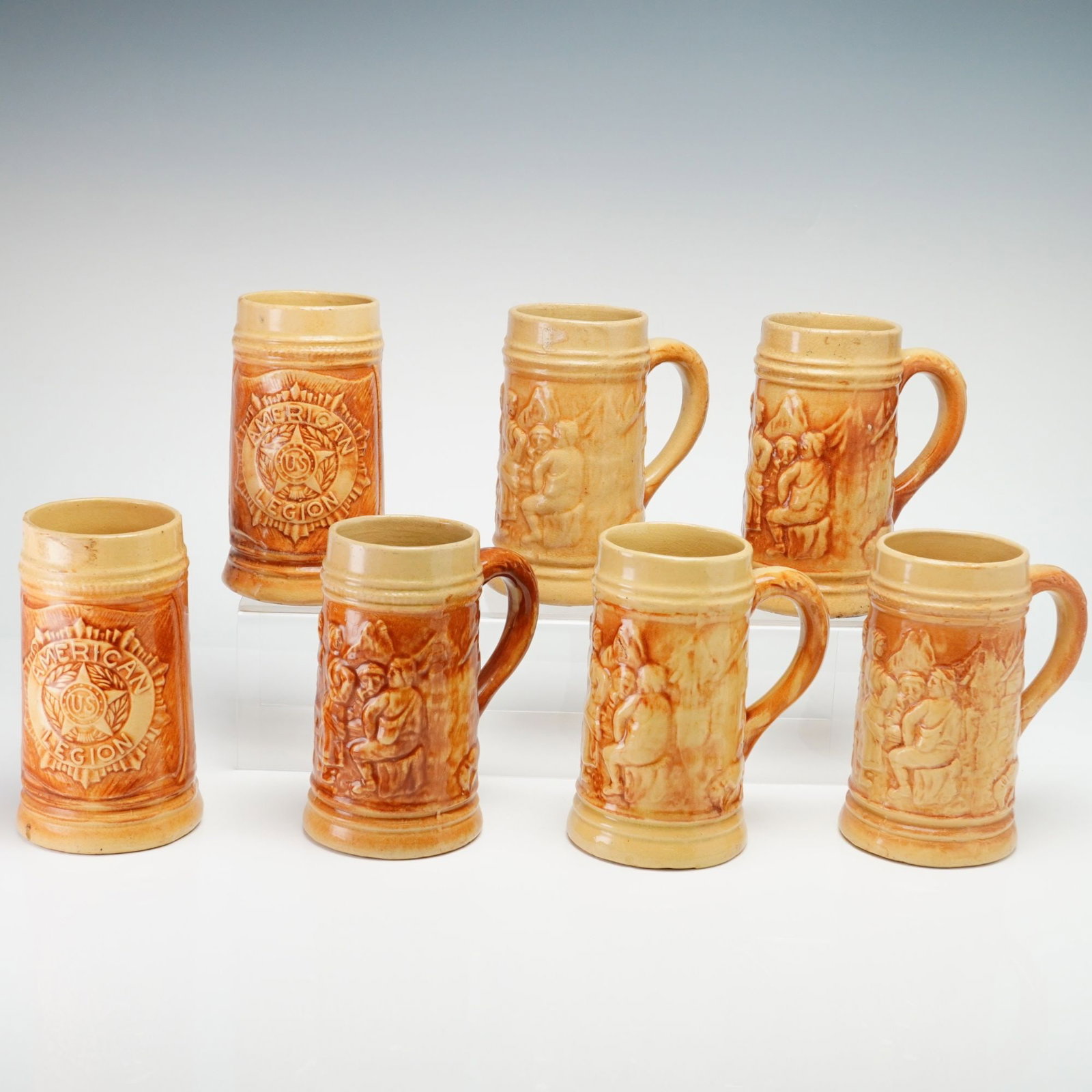 Set of Seven Hull Pottery Yellowware 1920s Steins, American Legion and Tavern Patterns: A set of seven stoneware mugs manufactured by Hull Pottery in Zanesville, Ohio. The collection features two distinct relief-molded designs. Two mugs display the American Legion emblem, which includes 