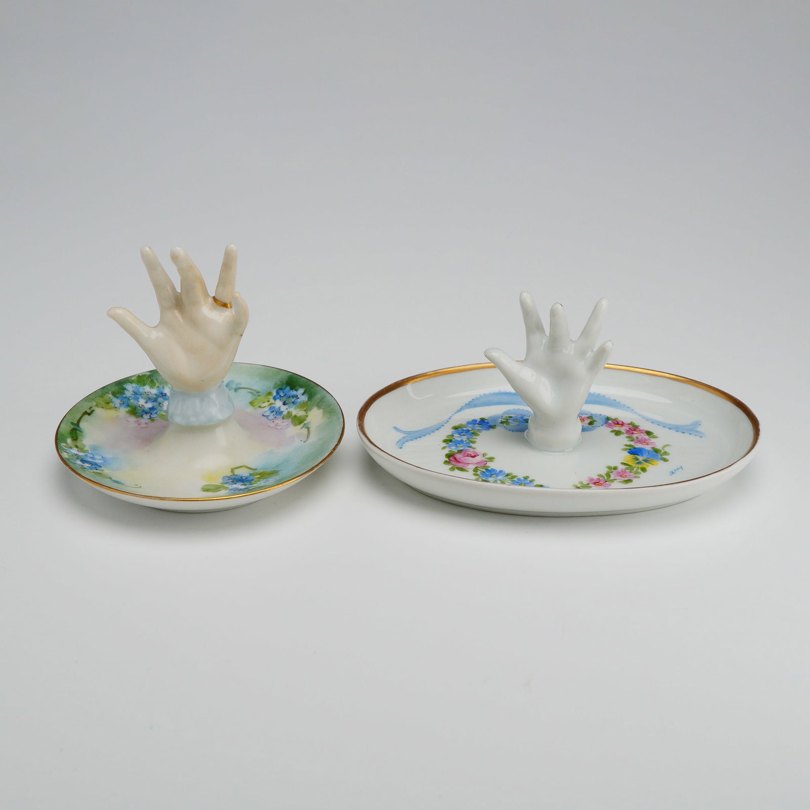 Two Limoges Porcelain Hand Form Ring Tree Dishes: A pair of vintage Limoges porcelain ring trees, each featuring a central upright hand-form post designed for jewelry display. The set includes one circular dish and one oval dish, both finished with h