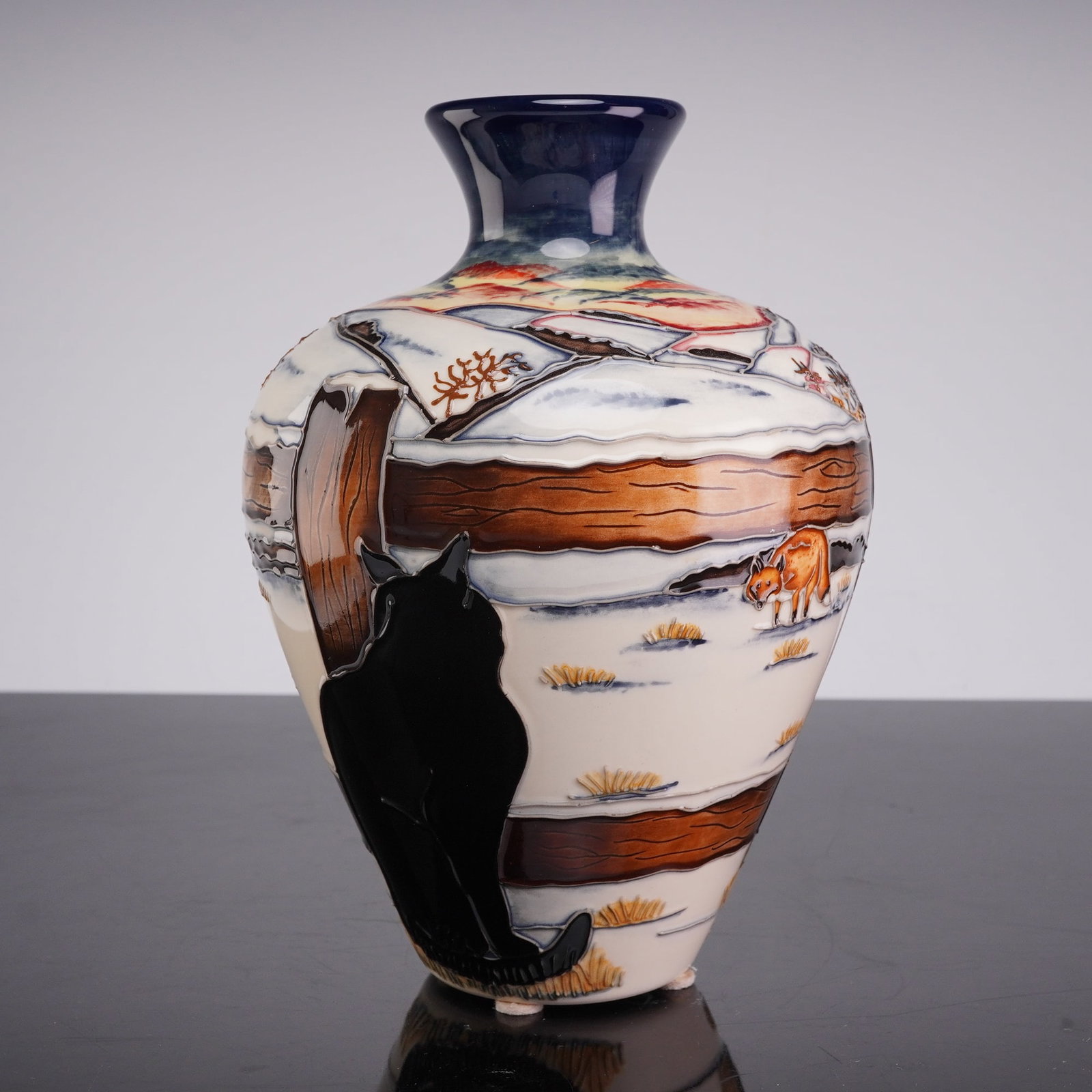 Moorcroft Pottery by Anji Davenport Vase, Woodside - Who Goes There?: This baluster-form vessel is decorated using a hand-painted tubelining technique, featuring a continuous winter landscape. The scene depicts a black cat in the foreground observing a fox across a snow