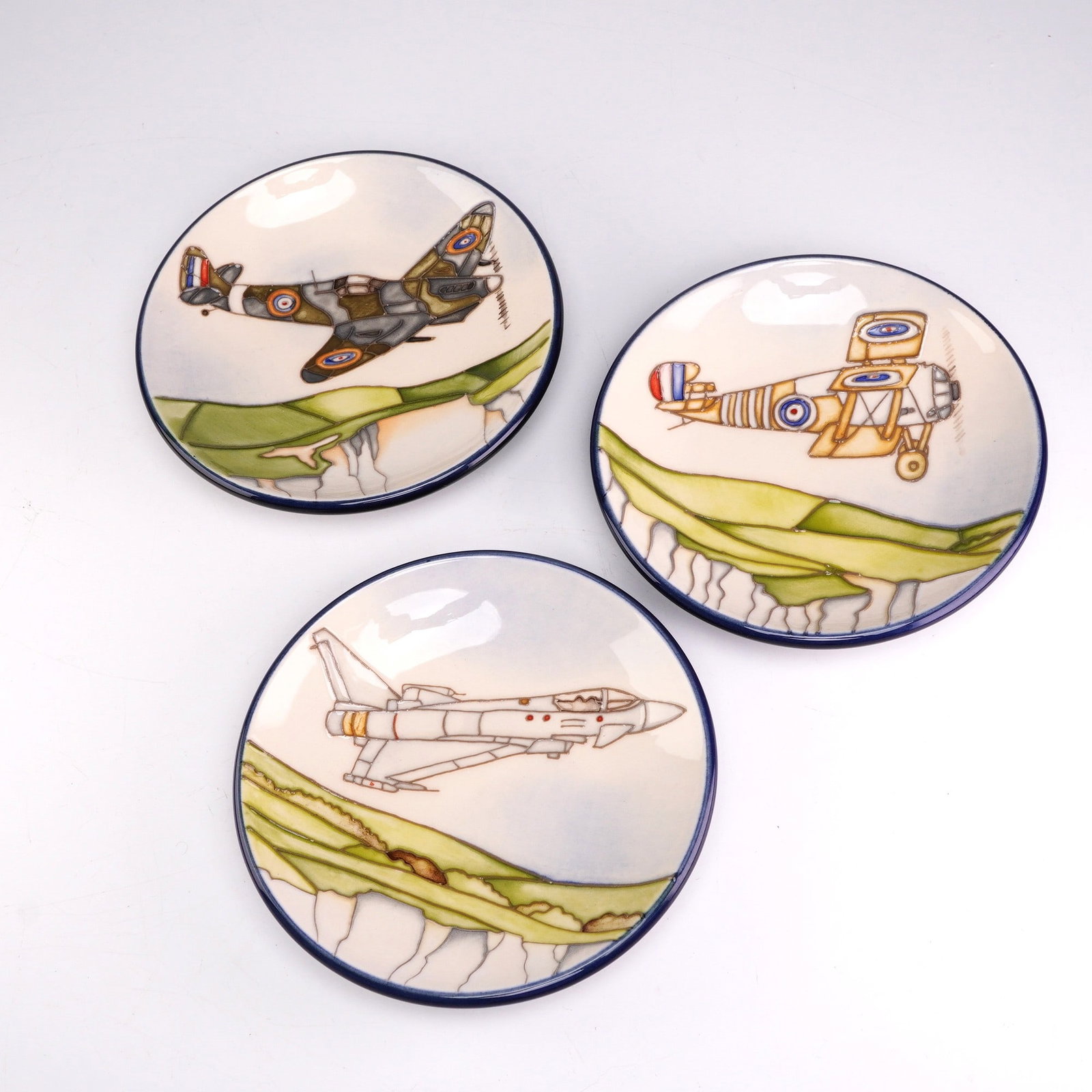 Three Moorcroft Pottery by Paul Hilditch Small Dishes, Flying Colours: This set of three small ceramic dishes was produced by Moorcroft Pottery as part of the Flying Colours series, designed by Paul Hilditch. Each circular dish features a distinct aviation-themed design 