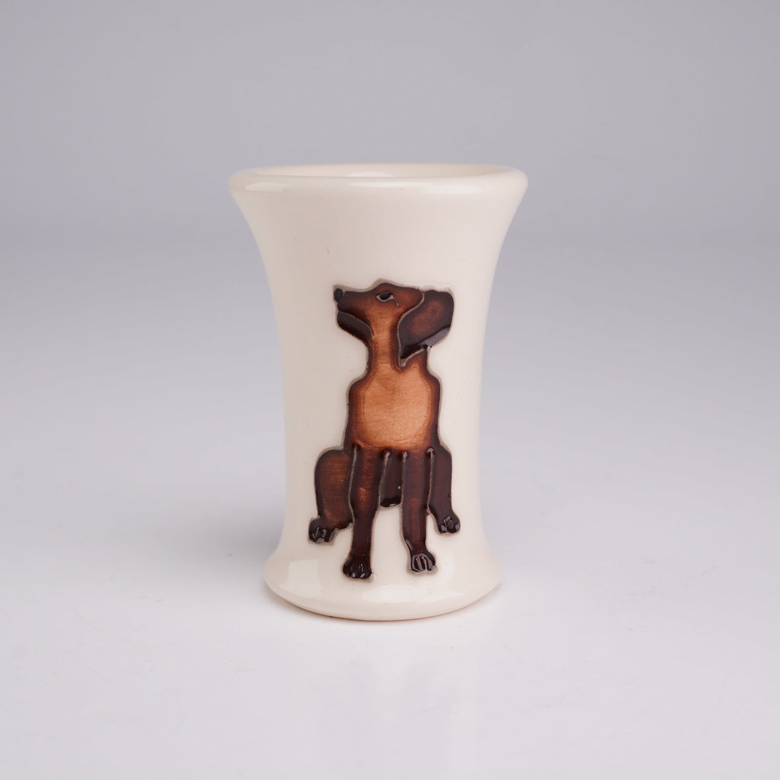 Moorcroft Miniature Dog TRIAL Vase: The piece features a waisted cylindrical form that flares at both the rim and the base. The decoration consists of two identical molded relief figures of a seated dog, possibly a dachshund or similar 