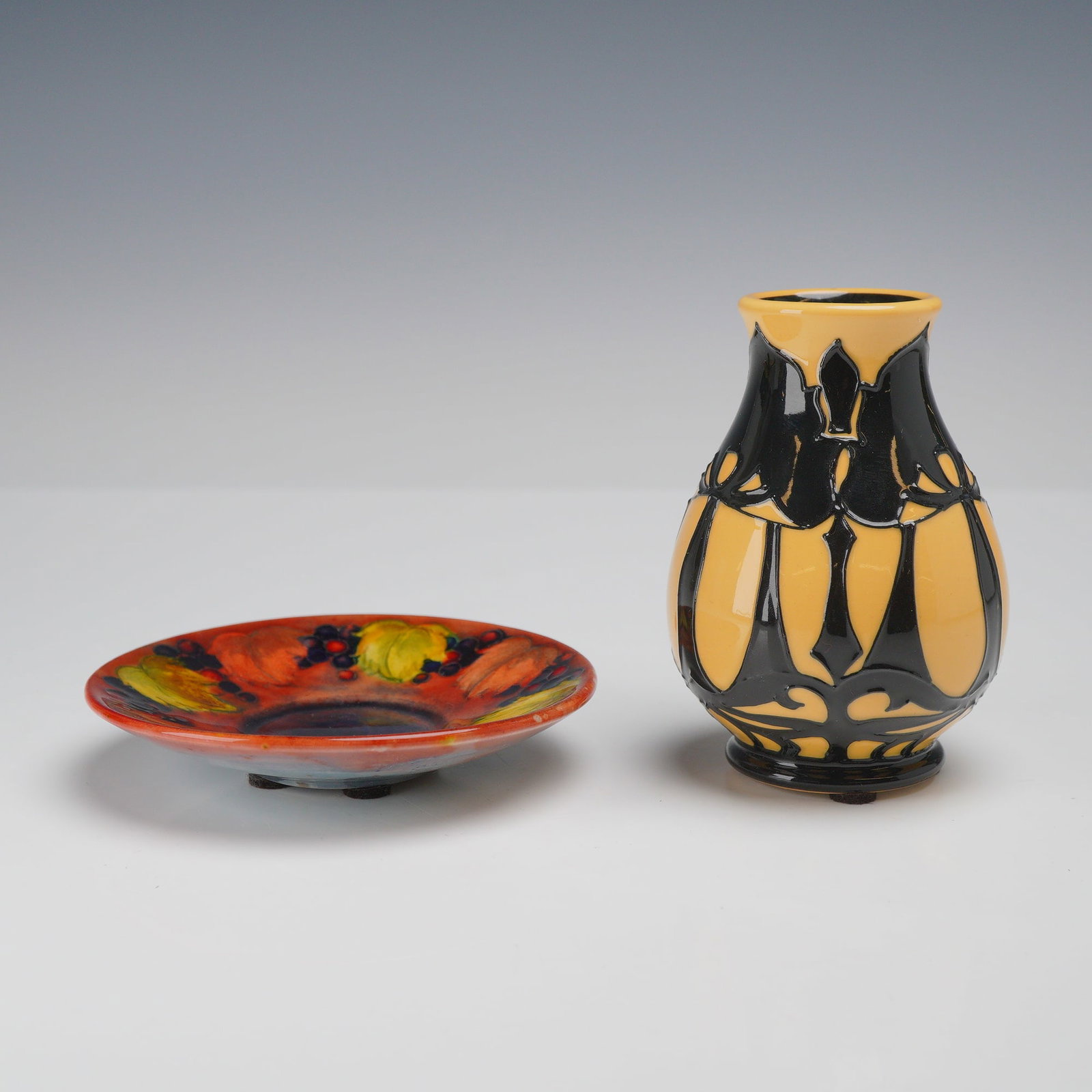 2pc Moorcroft Pottery Miniature Vase and Saucer Dish: This lot features two pieces of English art pottery from the Moorcroft factory. The first is a miniature saucer dish featuring a traditional fruit-themed pattern with tube-lined decoration depicting g