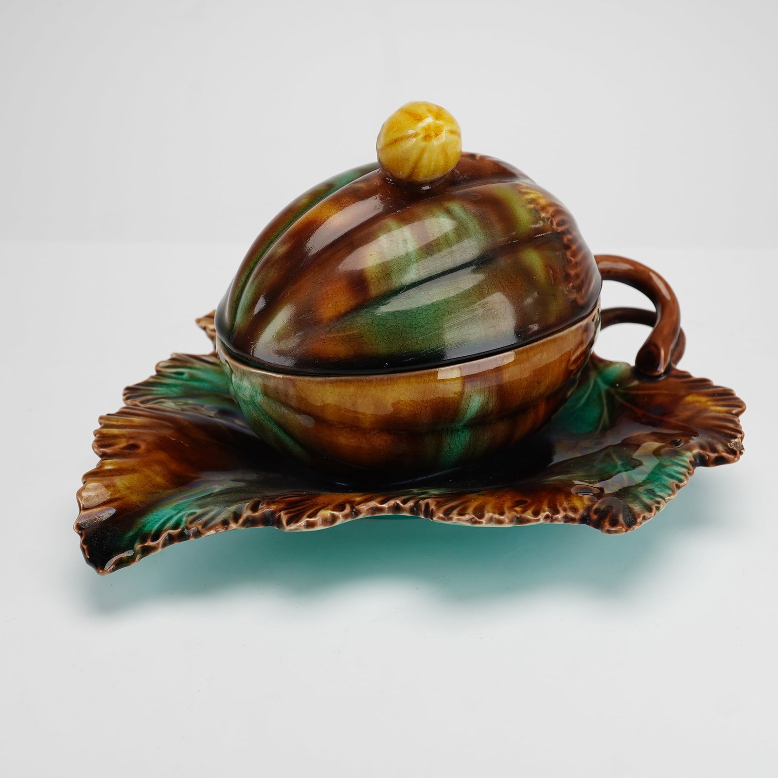 Wedgwood Majolica Tortoiseshell Glaze Lidded Condiment Pot on Leaf Stand: This Wedgwood majolica lot features a lidded ceramic condiment pot on a molded leaf-shaped stand. The vessel is designed in the form of a ribbed gourd or melon, topped with a bright yellow bud finial.