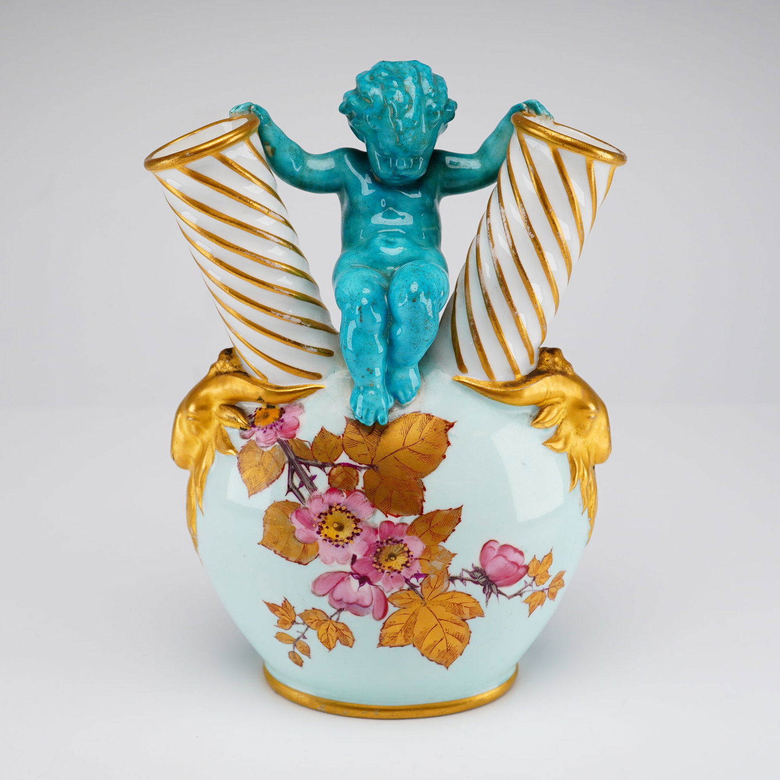 English Porcelain Double-Neck Putto Vase Attributed to Wedgwood: A porcelain double-neck vase, attributed to Wedgwood, designed with a central turquoise-glazed figure of a seated putto. The figure is positioned with outstretched arms grasping two diverging, spirall